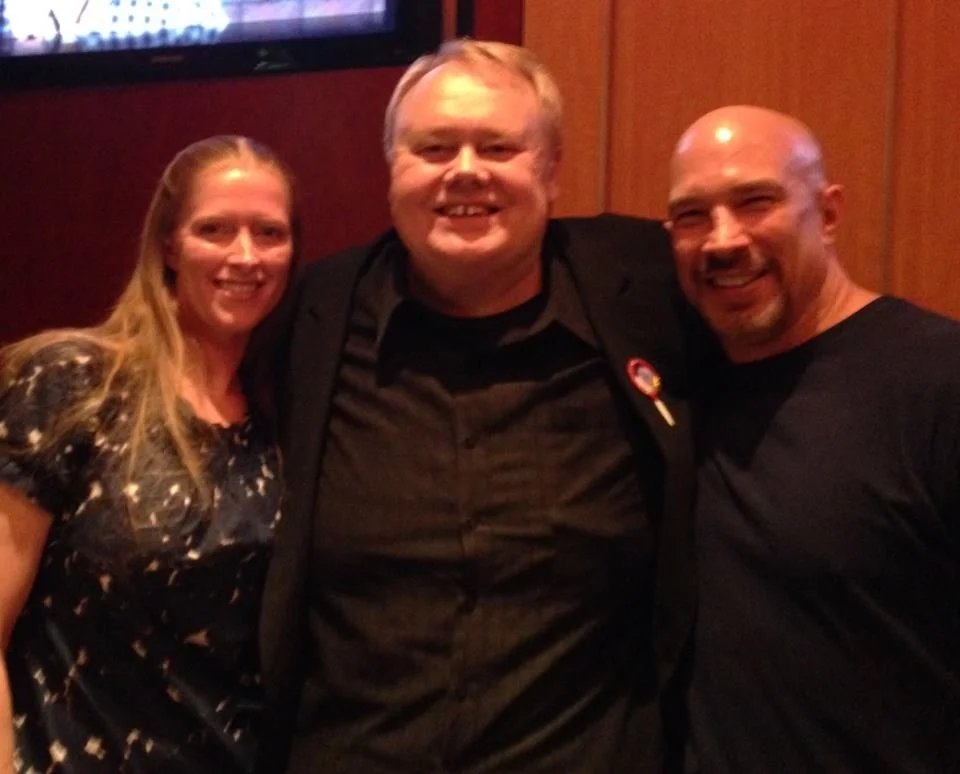 Vic Alejandro with his wife Amanda Alejandro and comedian Louie Anderson (RIP). Three people standing side by side, smiling, in a room with wood-paneled walls with Louie in the middle. 