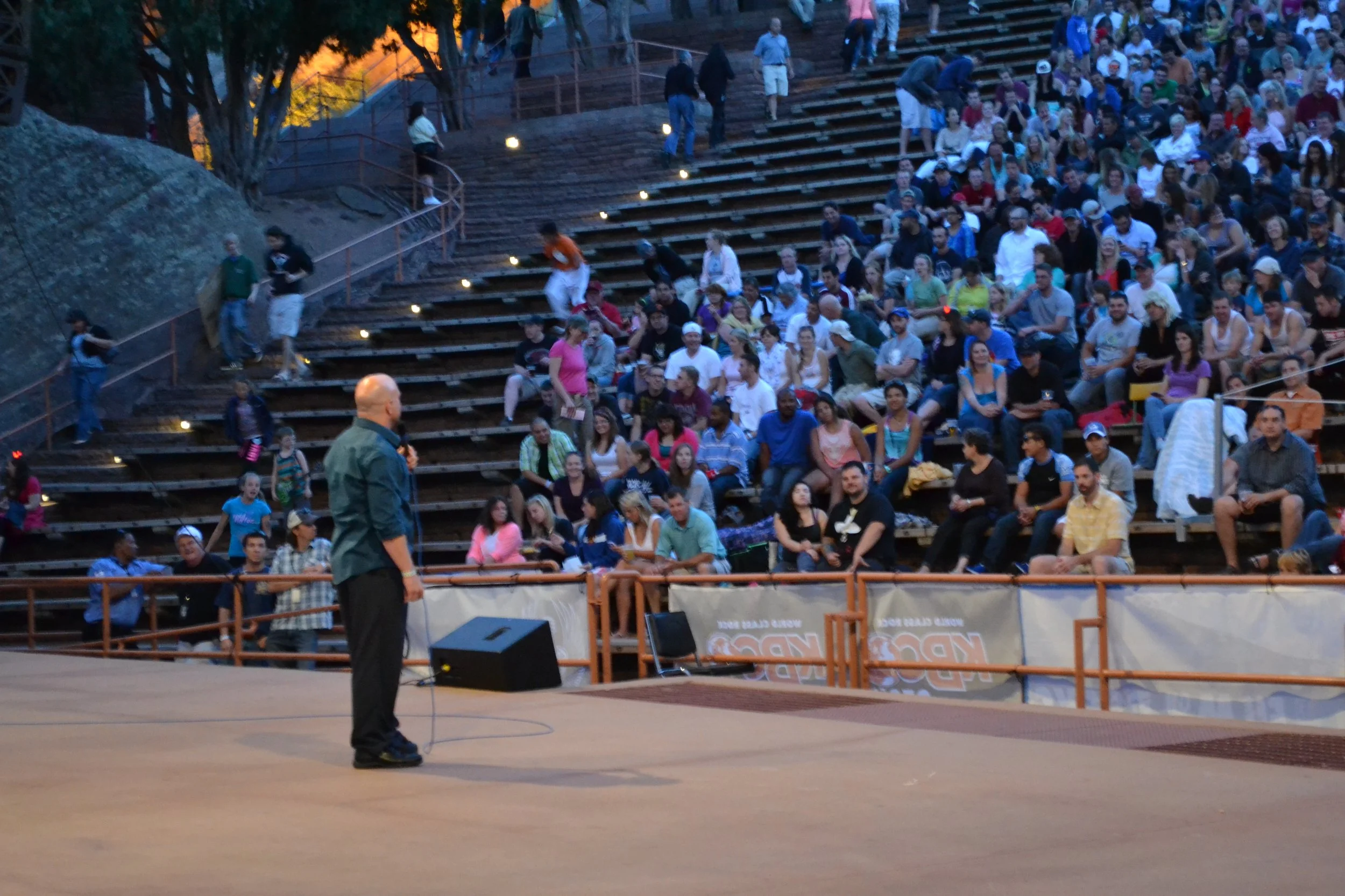 Vic Alejandro performing stand-up comedy at Red Rocks outdoor Amphitheatre in Colorado filled with a large crowd of people seated on tiered stone benches under a cliff face with lighting indicating early evening or dusk.