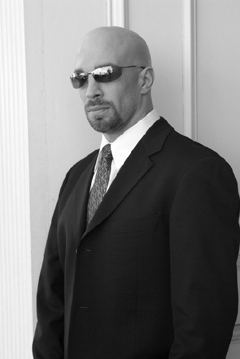 Vic Alejandro wearing sunglasses, a black suit, and tie standing against wall looking like a Secret Service agent.