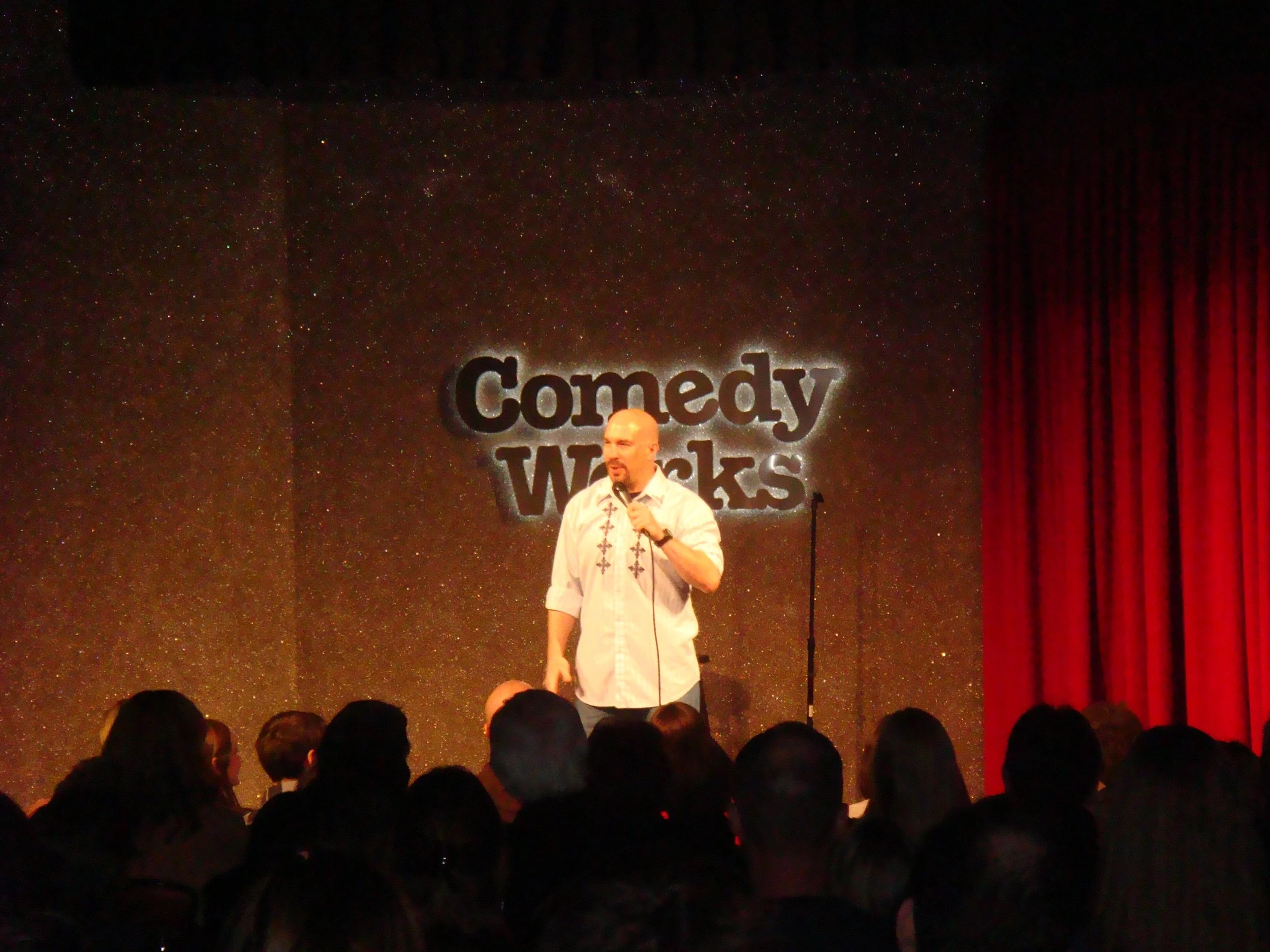 Vic Alejandro on stage at Comedy Works DTC Colorado. He stands on stage holding a microphone during a comedy show, with a large sign behind him reading "Comedy Works."