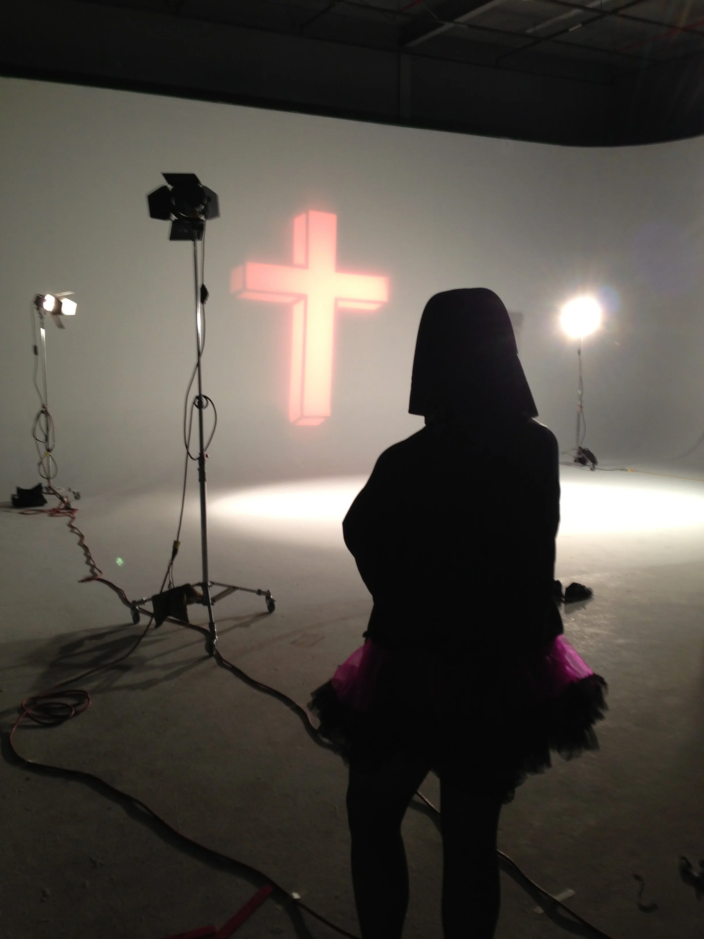 Vic Alejandro on the set of "Directors on Directing" dressed as a nun in a white habit and black tutu skirt standing in a studio with a large illuminated cross on the wall behind them. Studio lights and equipment are visible.