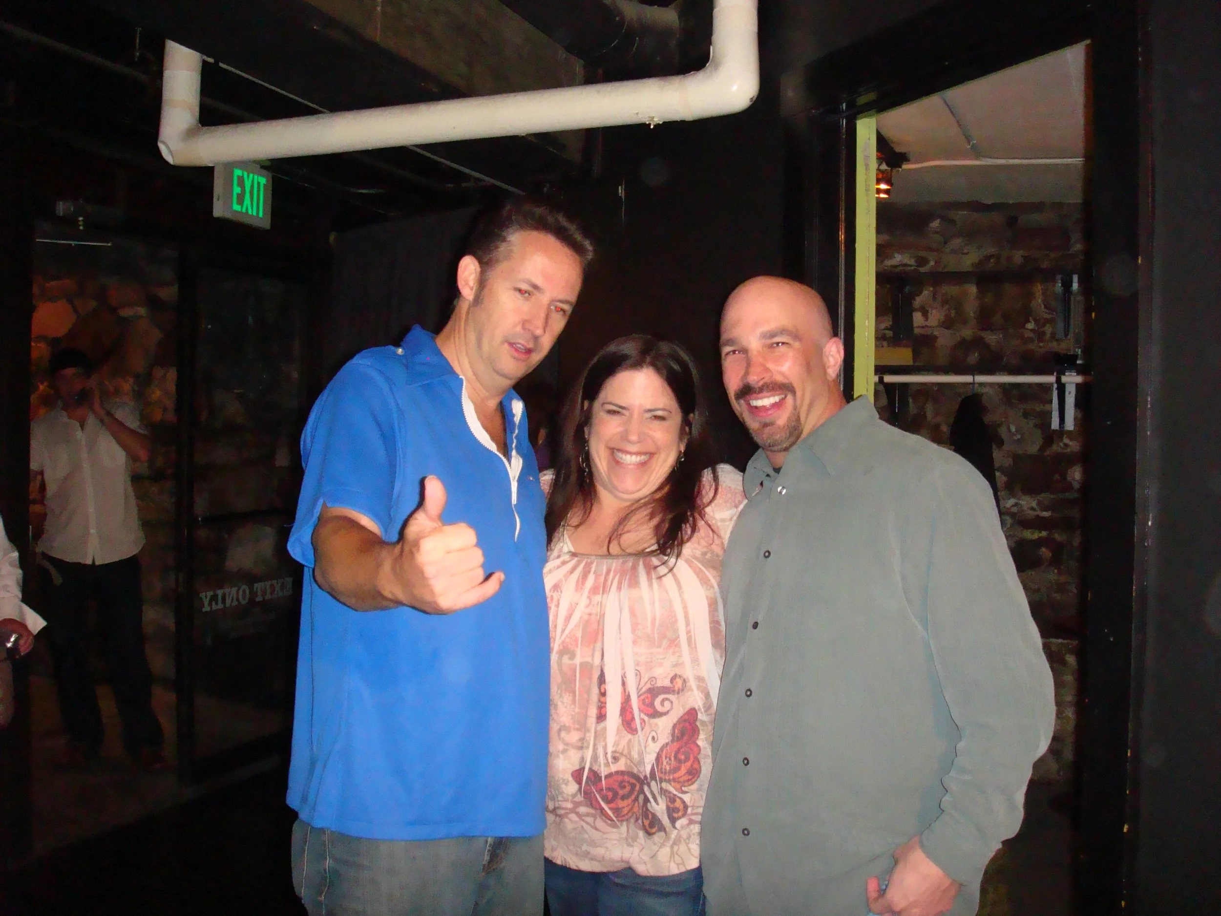 Vic Alejandro with comedians Harland Williams and Marla Schultz in front of the green room of Comedy Works in downtown Denver, Colorado. They're smiling and posing for a photo in a dimly lit indoor setting, with a Harland giving a thumbs-up gesture.