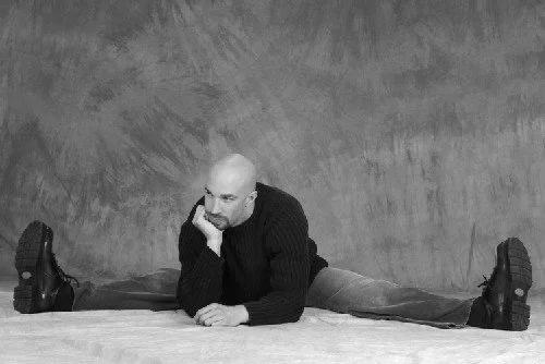 Vic Alejandro with a beard sitting on the floor with legs split wide apart, resting his head on his hand, against a plain textured background.