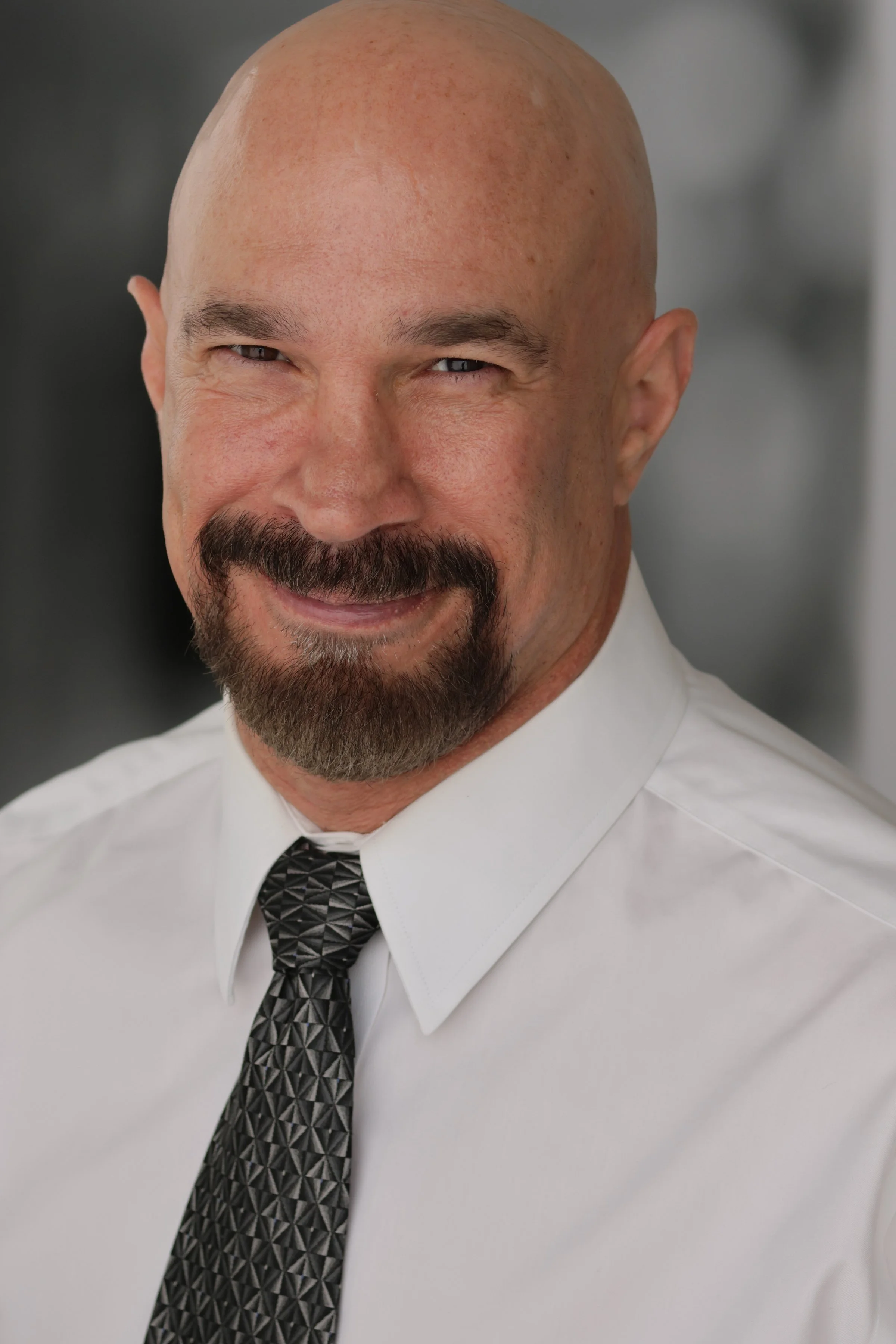 A close-up portrait of Vic Alejandro smiling with light skin, a bald head, and a neatly groomed beard and mustache. He is dressed in a white dress shirt and a patterned black and gray tie.