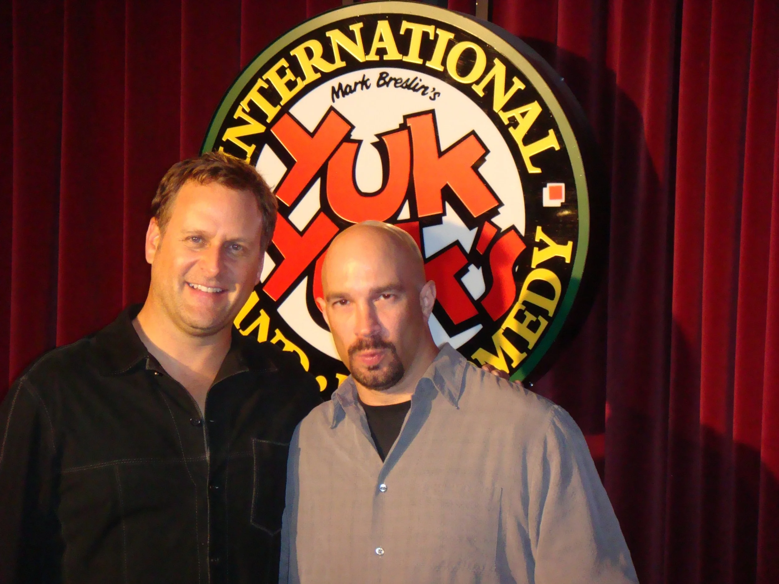 Vic Alejandro and Dave Coulier smiling in front of a circular sign that reads "Yuk Yuks" with a red and white design, red curtains in the background.