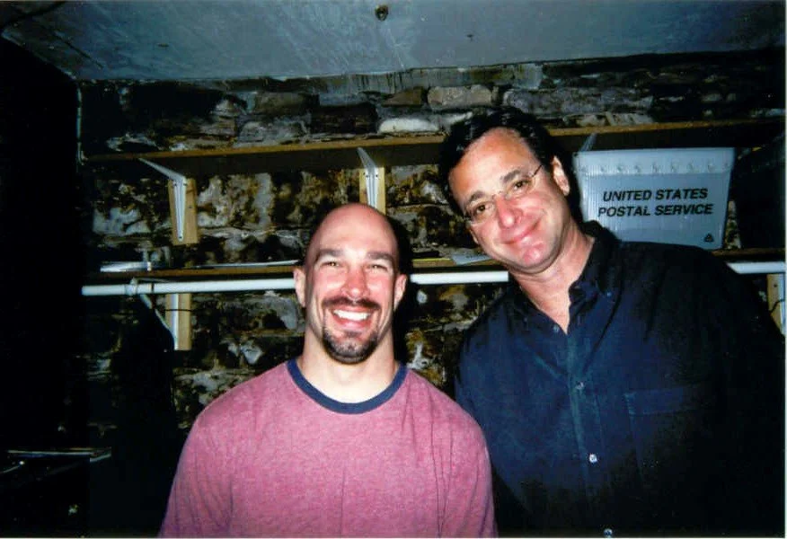 Vic Alejandro with comedian Bob Saget (RIP) in the green room of Comedy Works in downtown Denver, Colorado. Two men smiling in front of a stone wall. 