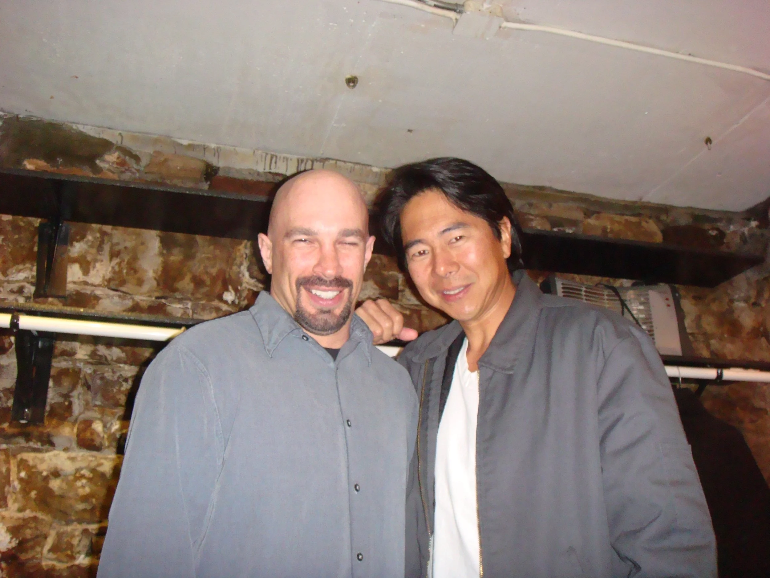 Vic Alejandro with comedian Henry Cho in the green room of Comedy Works in Denver. Two men are smiling and posing for a photo in front of a brick wall. They are dressed casually, one with a gray shirt and the other with a gray jacket and white shirt.