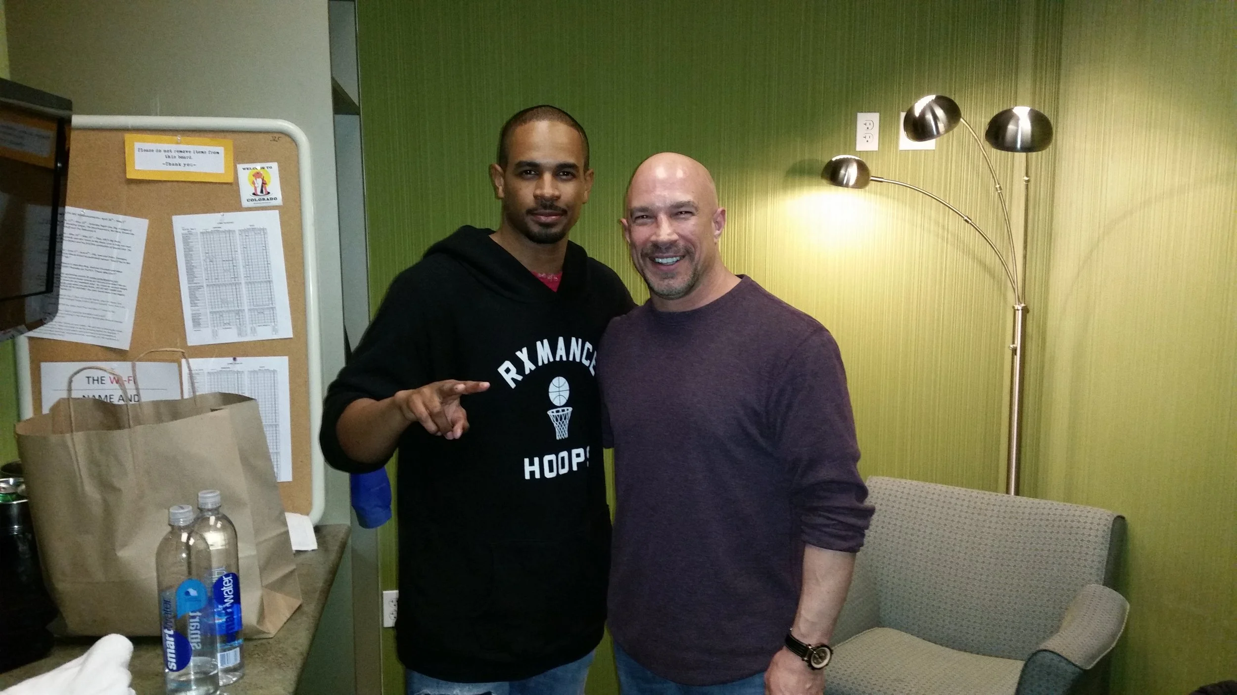 Vic Alejandro with comedian Damon Wayans Jr. in the green room at Comedy Works in DTC Colorado. Two men smiling while posing for a photo indoors with green walls, a lamp, a corkboard with papers, and bottles of water on a table.
