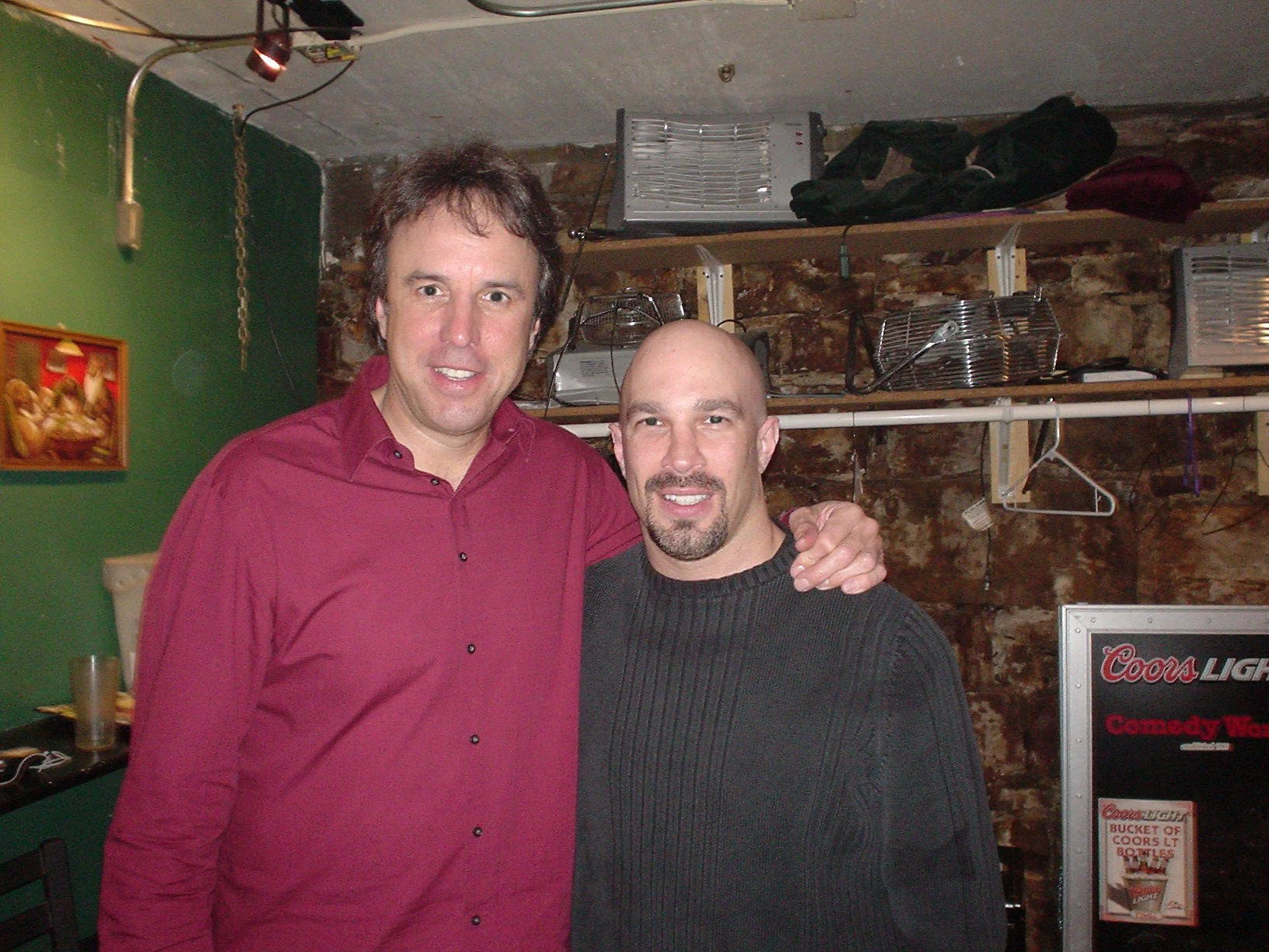 Vic Alejandro with comedian Kevin Nealon in the green room of Comedy Works in Denver standing close together, smiling at the camera. The background reveals a green wall and a brick wall with shelves