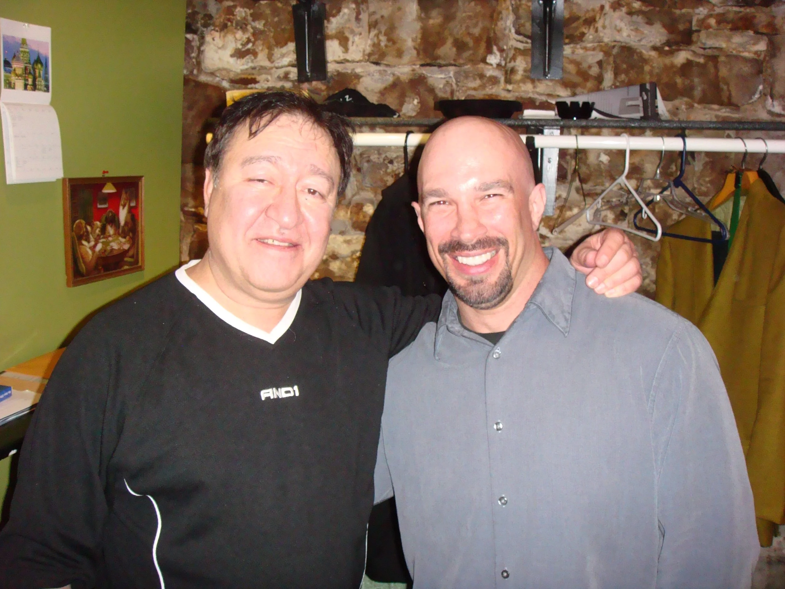 Vic Alejandro with comedian Dom Irerra in the green room of Comedy Works in downtown Denver, Colorado. They stand close together in front of a brick wall with a green wall and framed pictures in the background.