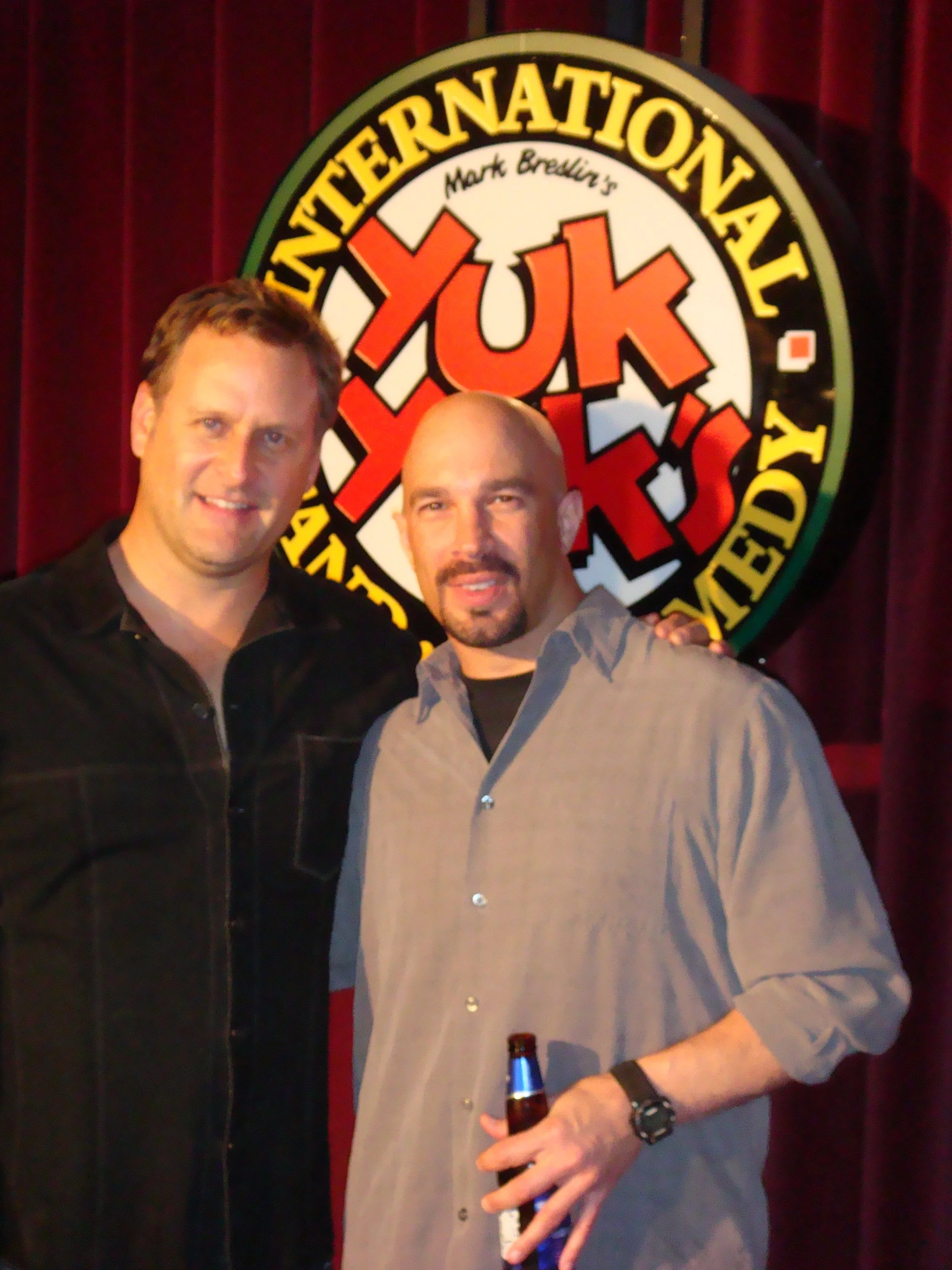 Vic Alejandro and Dave Coulier smiling in front of a circular sign that reads "Yuk Yuks" with a red and white design, red curtains in the background. Vic has a beer in his hand.