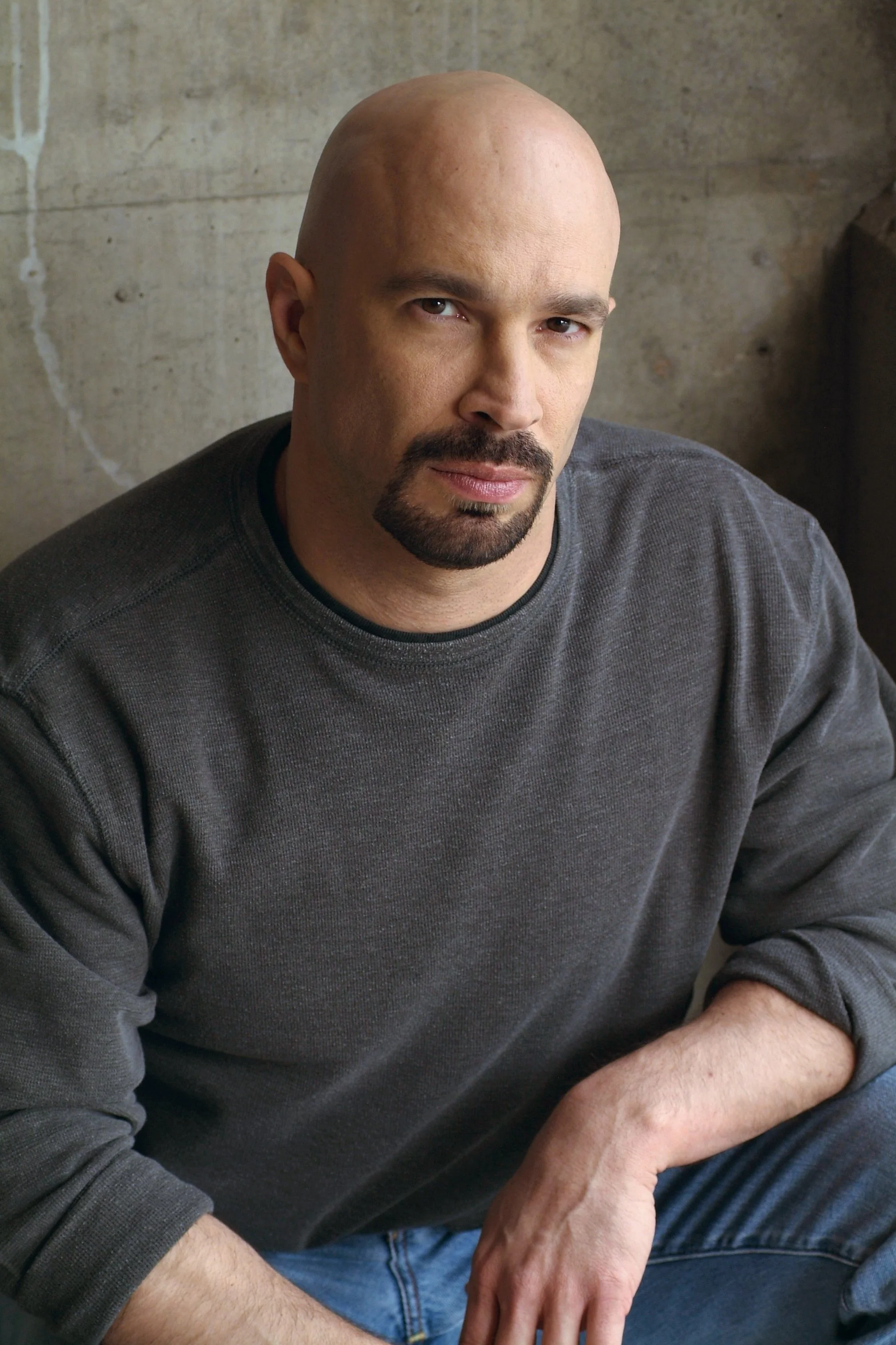 Vic Alejandro with a shaved head and goatee, wearing a gray sweatshirt, sitting against a concrete wall.