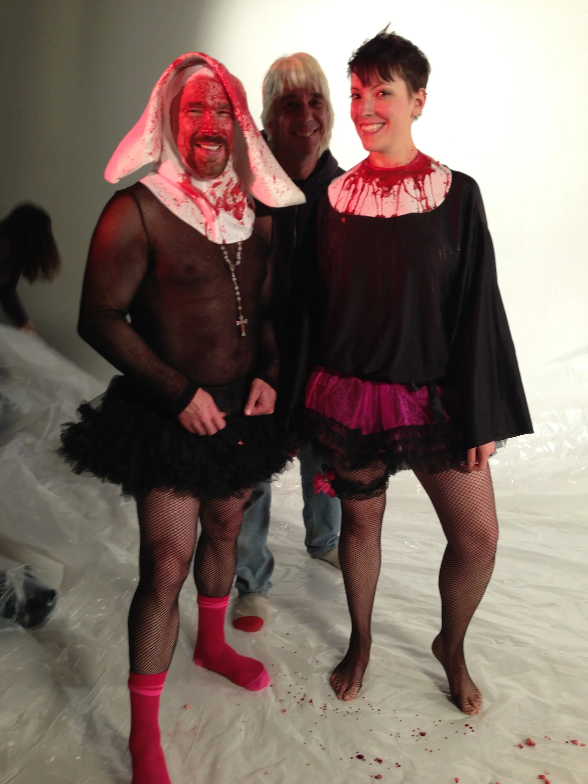 Vic Alejandro on the set of "Directors on Directing" dressed as a nun in a white habit and black tutu skirt standing in a studio with a female co-star with short black hair in a similar outfit and Director Damien with short bleached hair. Wide shot.