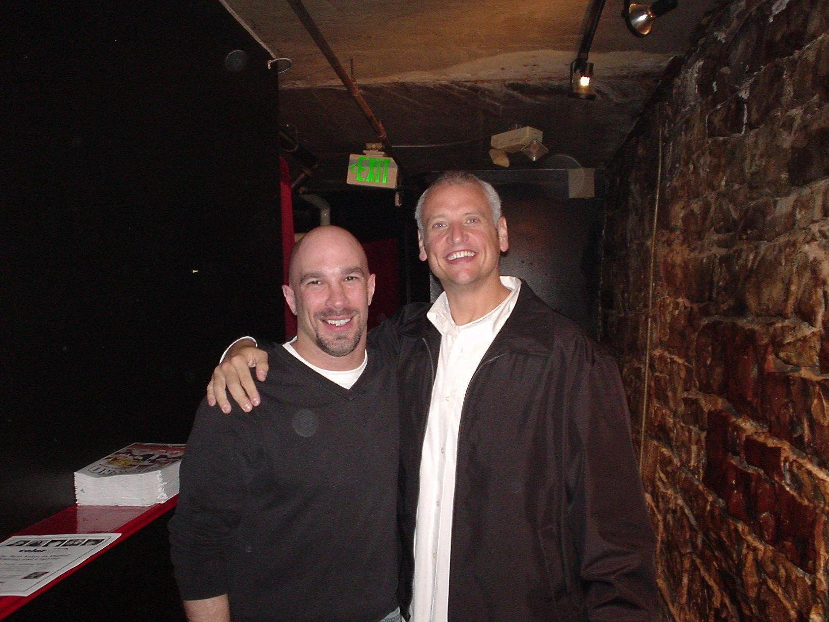 Vic Alejandro with comedian Eddie Gosling in front of the green room of Comedy Works in downtown Denver, Colorado with their arms around each other's shoulders against a brick wall.