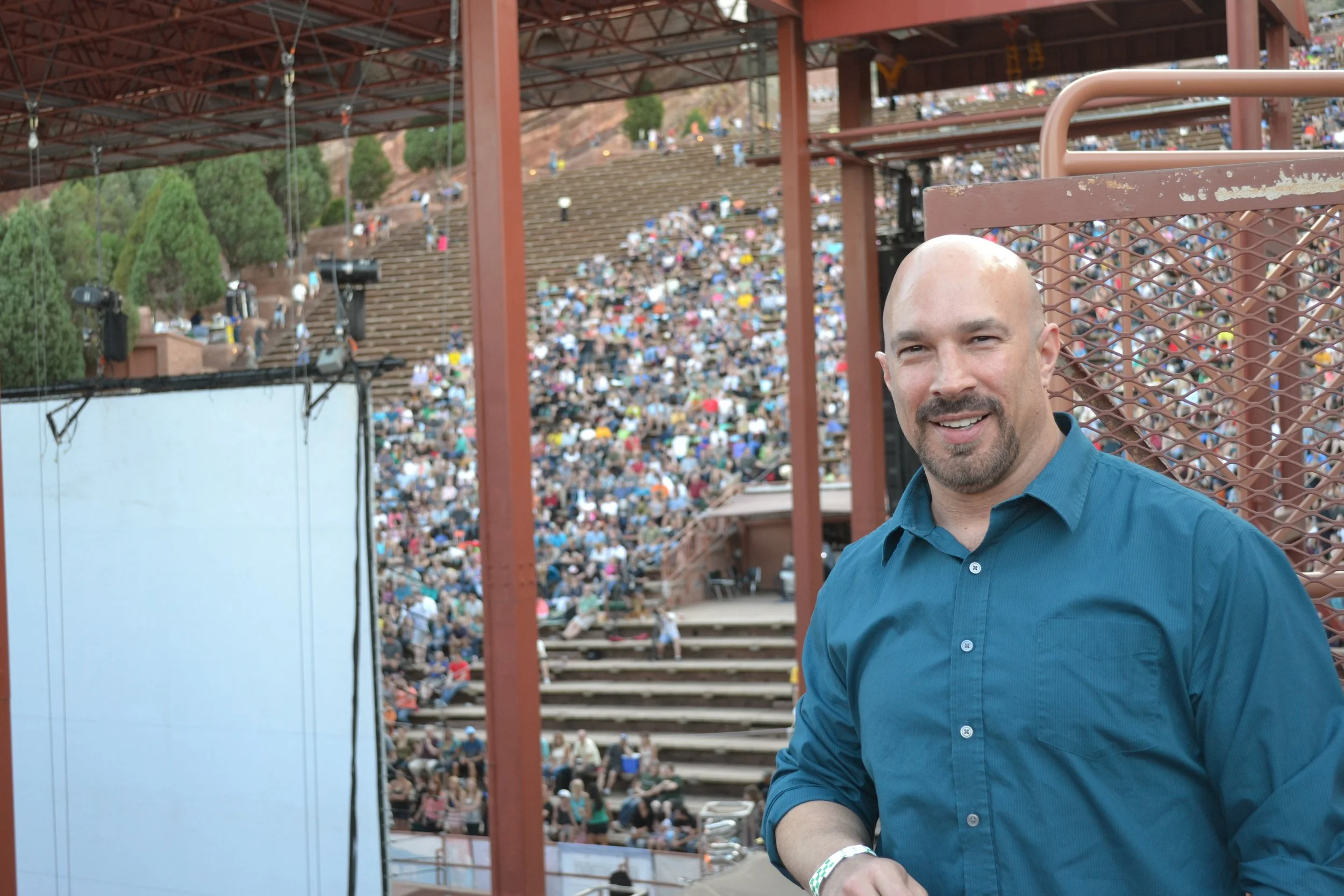 Vic Alejandro from an elevated position backstage at Red Rocks outdoor Amphitheatre in Colorado with a large crowd seated in tiered rows in the background.