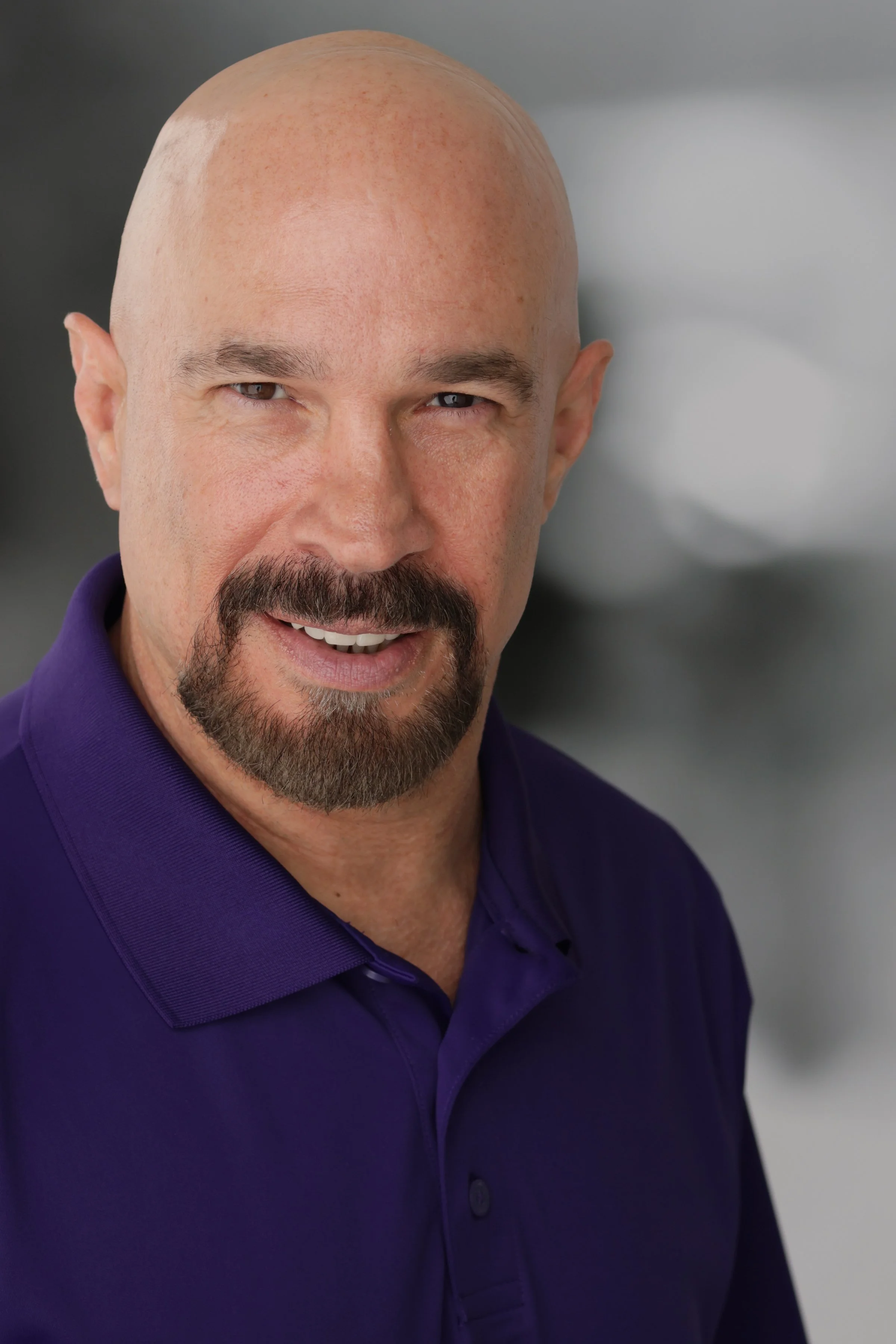 Vic Alejandro smiling with a mustache and beard, bald head, wearing a purple polo shirt, against a neutral background