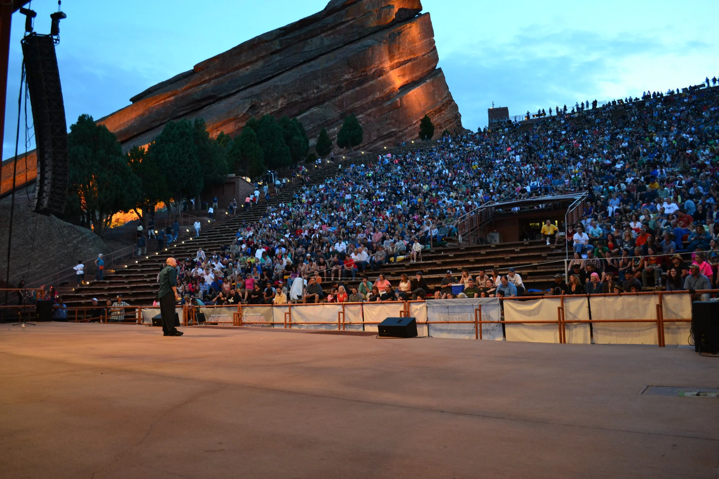 Vic Alejandro on stage at Red Rocks amphitheater in Colorado filled with a large audience seated with a mountain formation illuminated in orange in the background, and trees between the audience and the formation, during twilight.