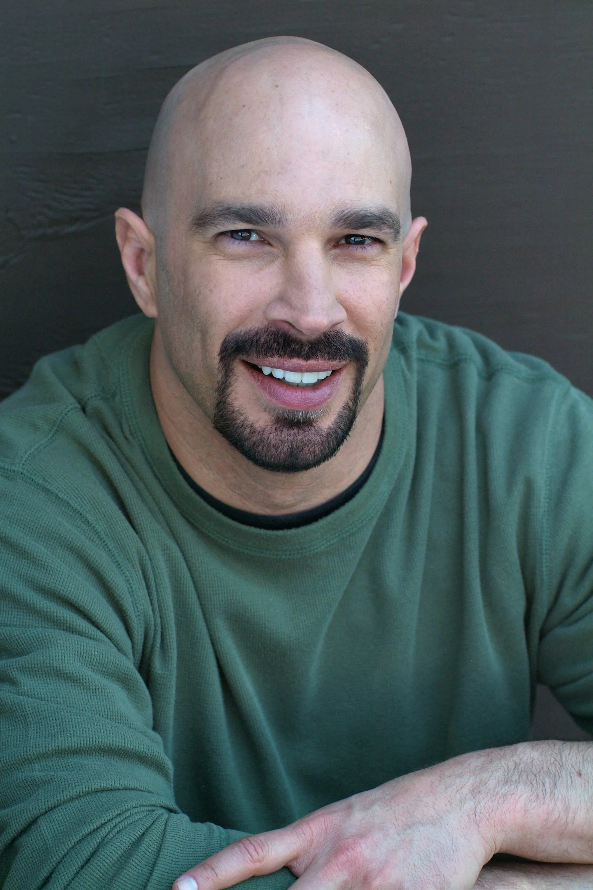 Vic Alejandro with a mustache and beard wearing a green shirt, sitting at a table with his arm resting on it, against a dark wooden background.