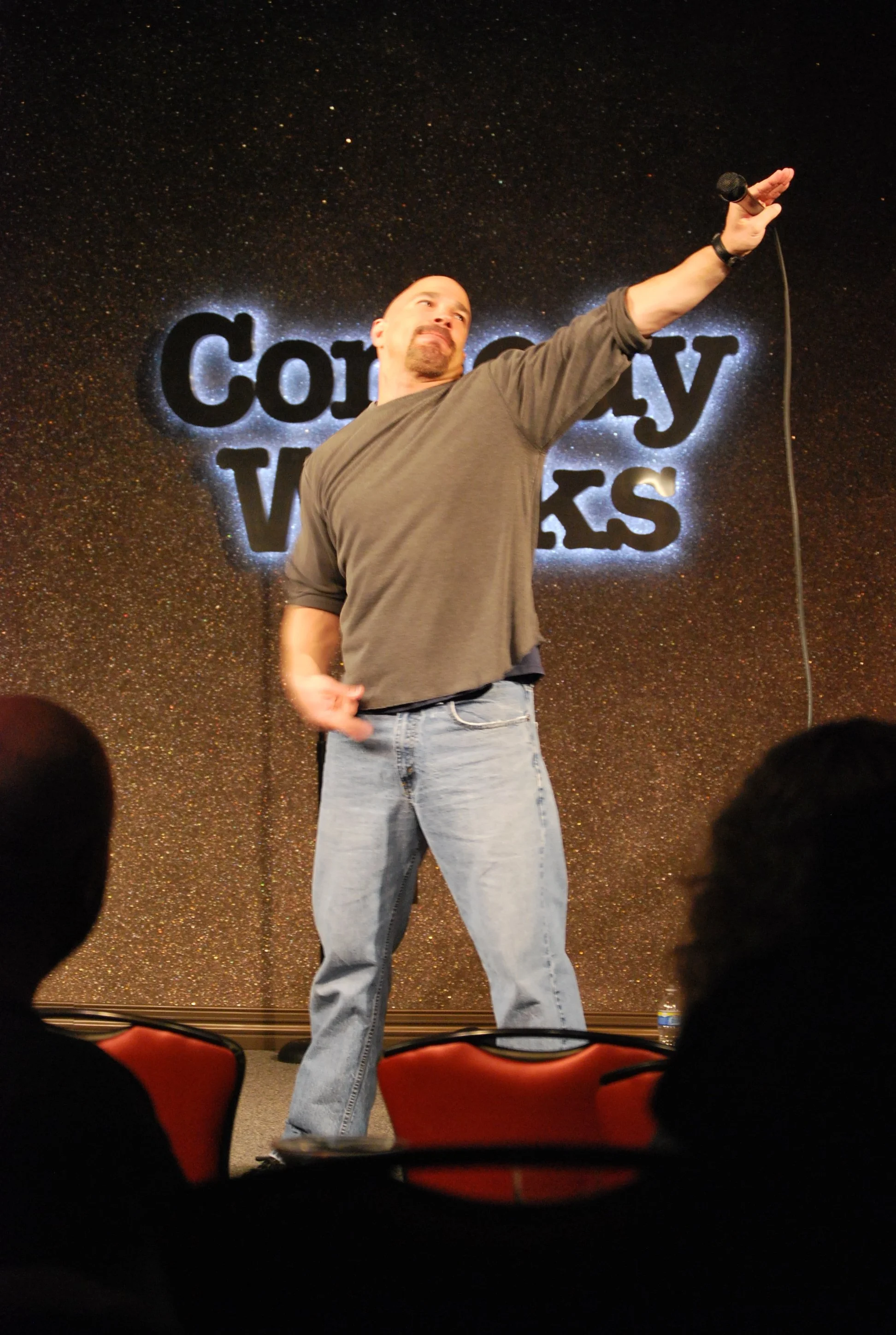 Vic Alejandro standing on a stage at Comedy Works in DTC holding a microphone in his right hand, with his left arm extended and pointing upwards. The background has a glittery texture with a sign that reads 'Comedy Works' partly visible behind him.