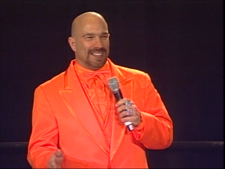 Vic Alejandro in an orange tuxedo, standing on a stage with a black backdrop, speaking into a microphone at the PMI Conference in Denver, Colorado.