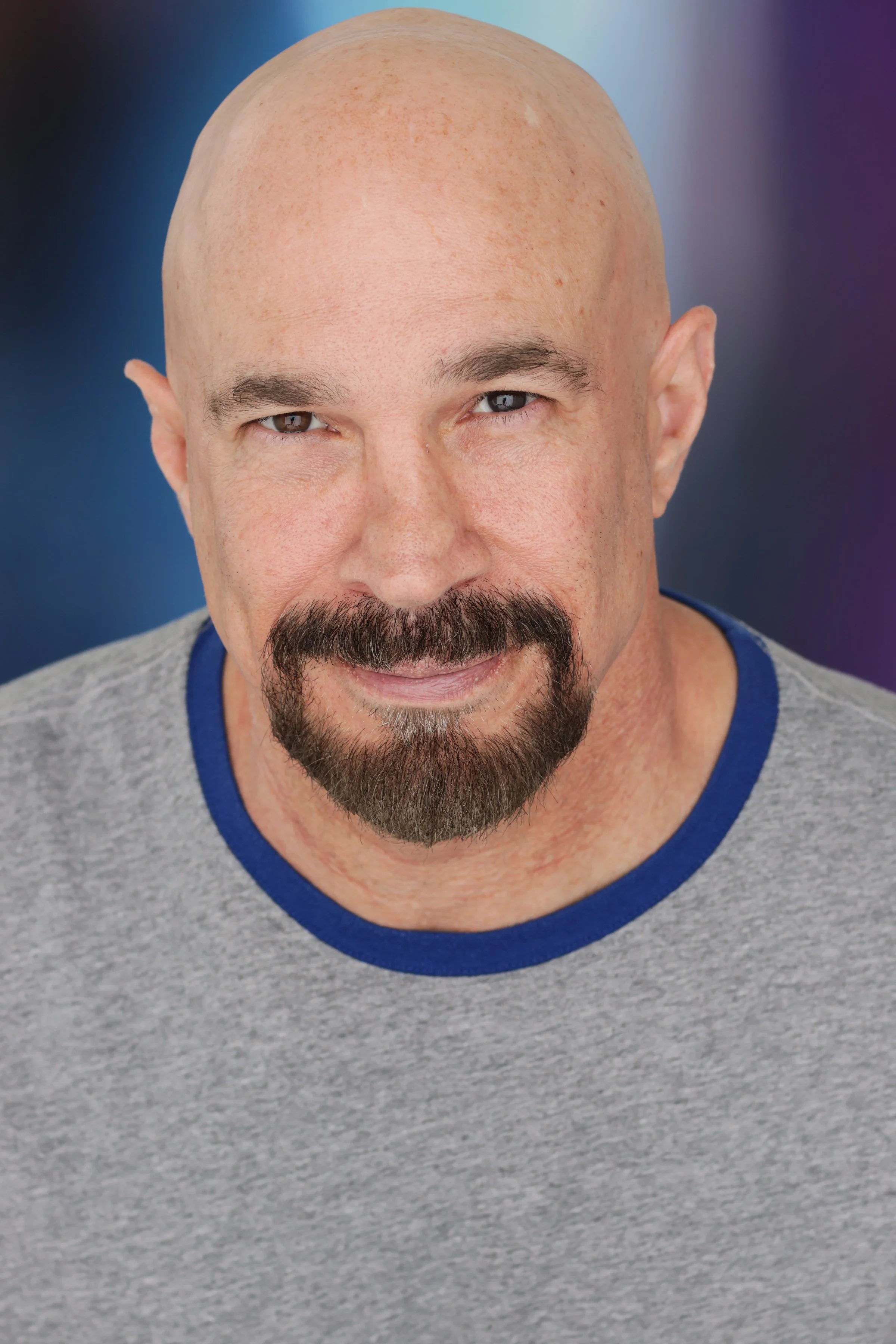 Close-up of Vic Alejandro. He is bald man with a goatee and mustache wearing a gray shirt with blue trim, against a blurred blue and purple background.