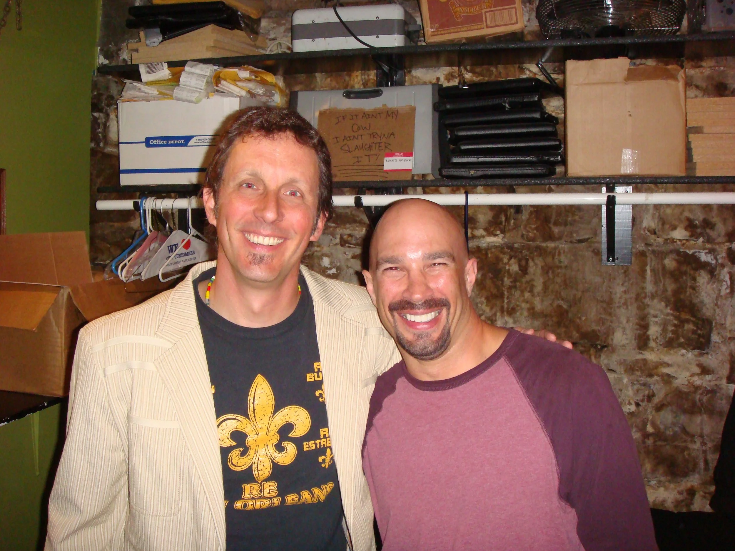 Vic Alejandro with comedian Jake Johannsen in the green room of Comedy Works in downtown Denver, Colorado. Two smiling men standing close together with shelves, boxes, and miscellaneous items in the background.