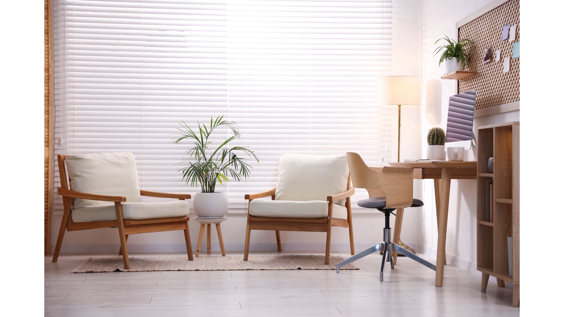 A well light room with two cream upholstered chairs facing each other.  A white potted plant between the two chairs with a lit corner lamp.  A wooden swivel chair at wooden desk with a desktop computer, cactus plant and mug sits on it.