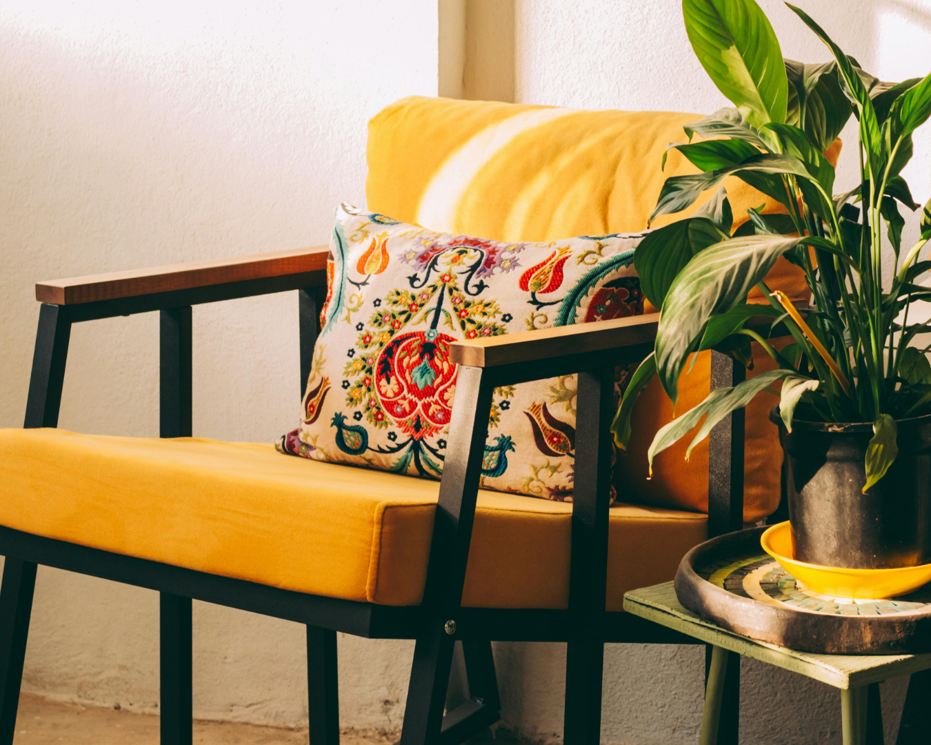 A cozy corner with a yellow cushion couch, a decorative embroidered pillow, a green potted plant, a small side table, and a light-colored wall in the background.