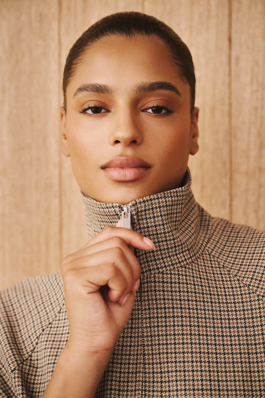 A woman with dark hair pulled back, wearing a houndstooth patterned zip-up jacket, standing in front of a wooden wall.
