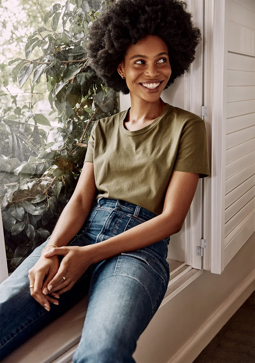 A woman with curly hair sitting by a window, smiling and looking to her left, wearing a green t-shirt and blue jeans.