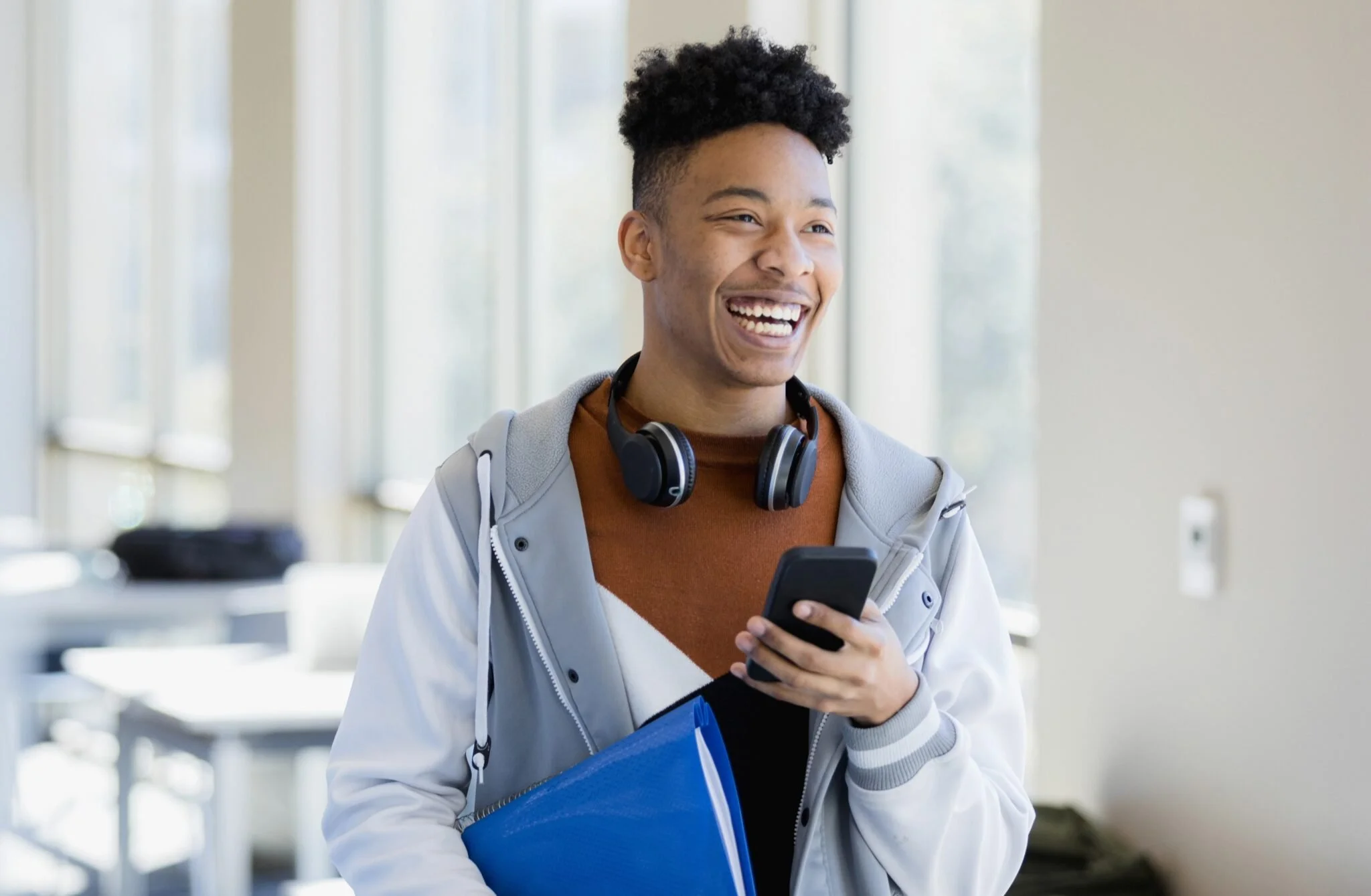 Young man with headphones around his neck holding a smartphone and a blue folder, smiling in a bright indoor setting.