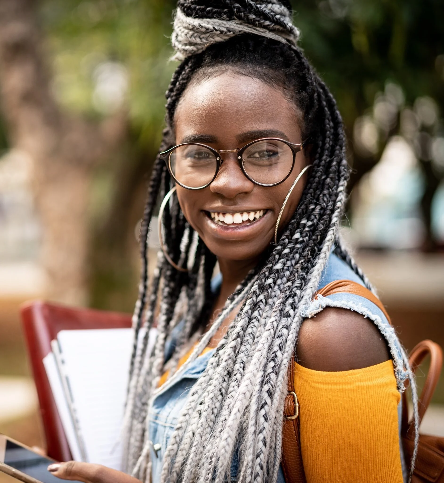 Close-up of a young woman with glasses smiling, with braided hair and wearing a yellow shirt, outdoors in a park.
