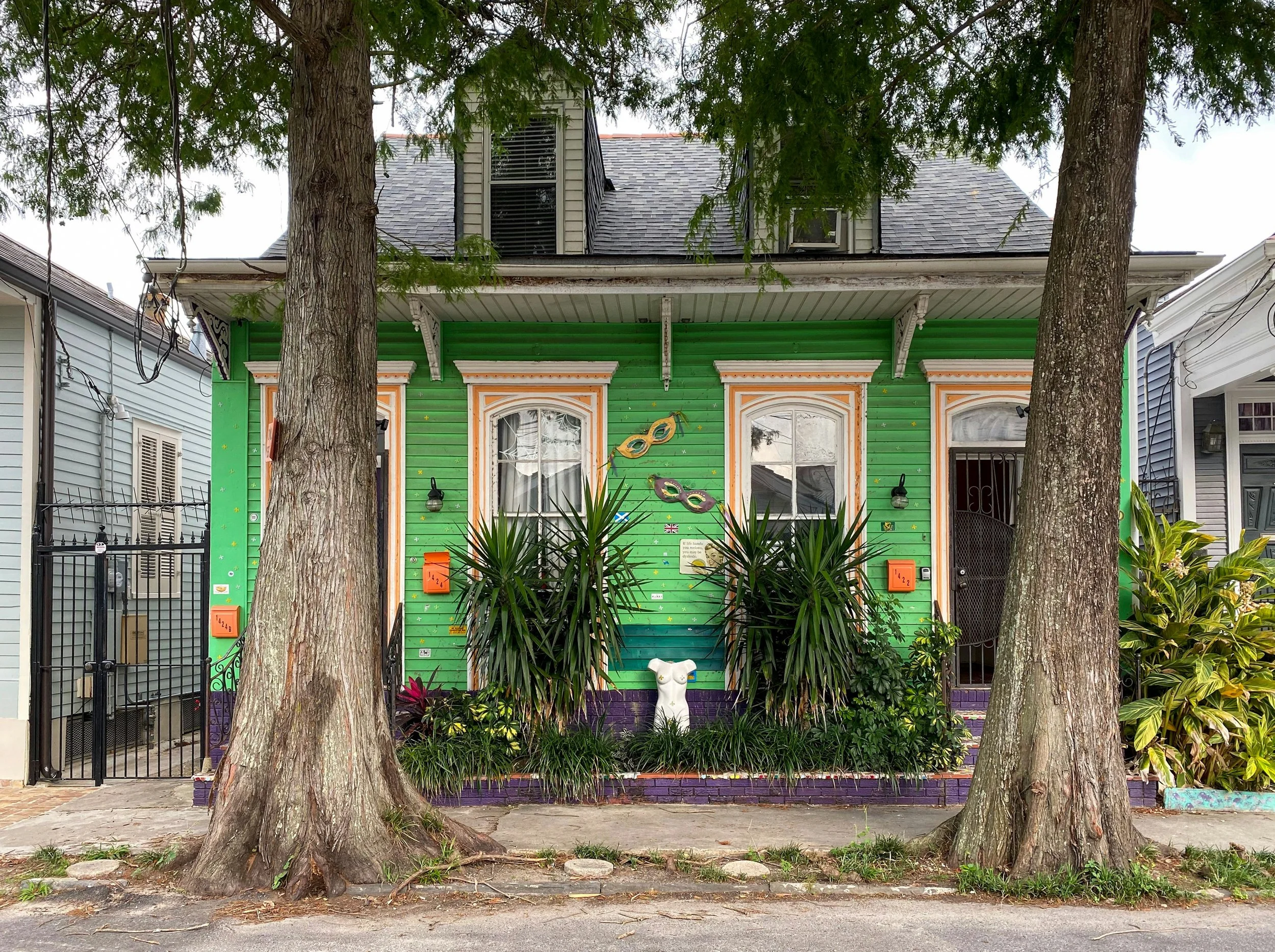 A colorful house painted in green, purple, and orange with decorative masks on the front wall, surrounded by trees and plants, with a dog statue in front.