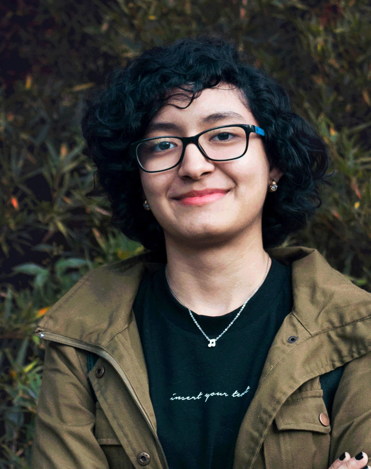 A young woman with curly black hair, glasses, and earrings smiling outdoors, wearing a black t-shirt, a brown jacket, and a silver necklace with a musical note pendant.