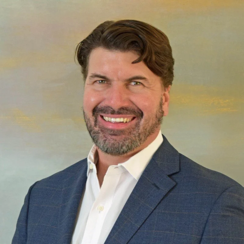 A smiling man with dark brown hair and a beard wearing a navy blue suit and white shirt, standing against a soft, abstract background.