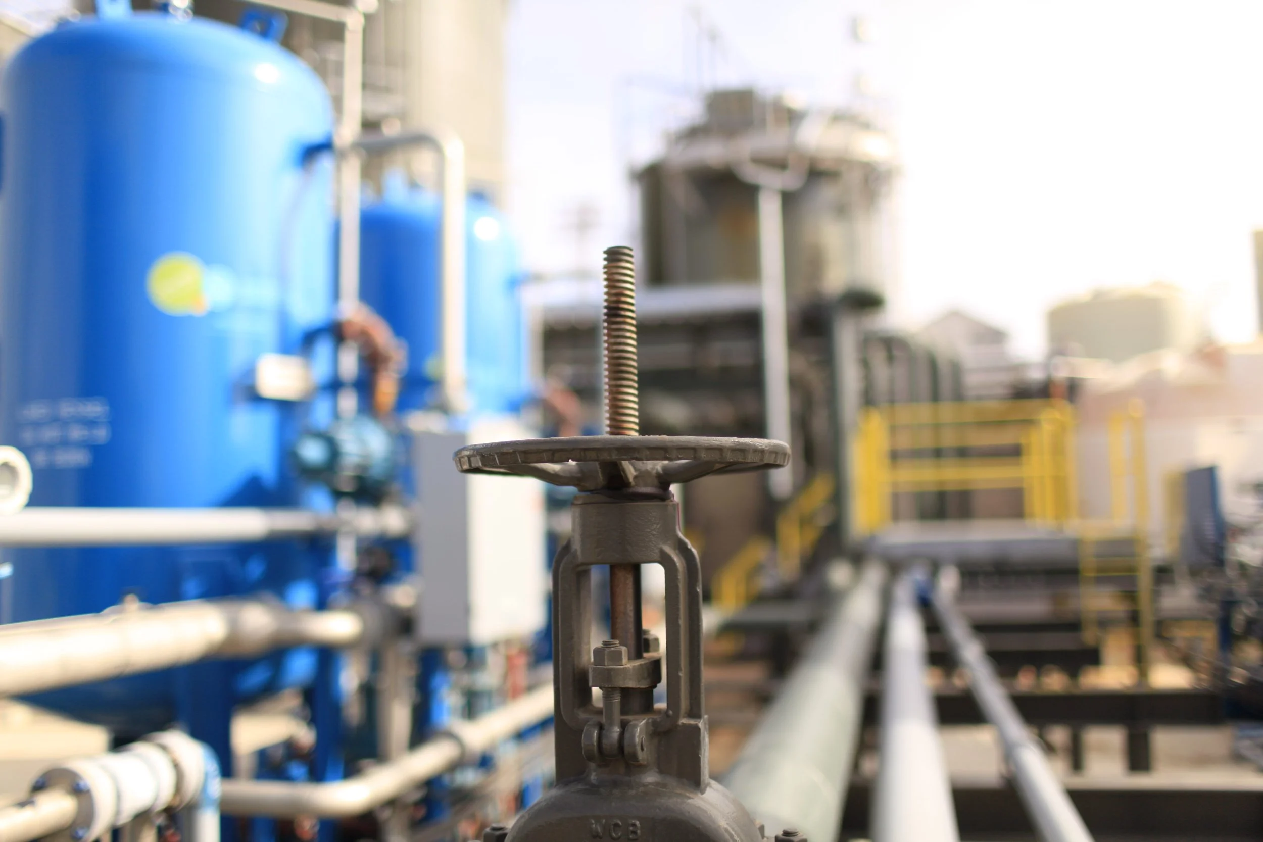 A close-up of a metal pipe fitting with a threaded pipe standing upright, in an industrial setting with blue tanks and pipes in the background.