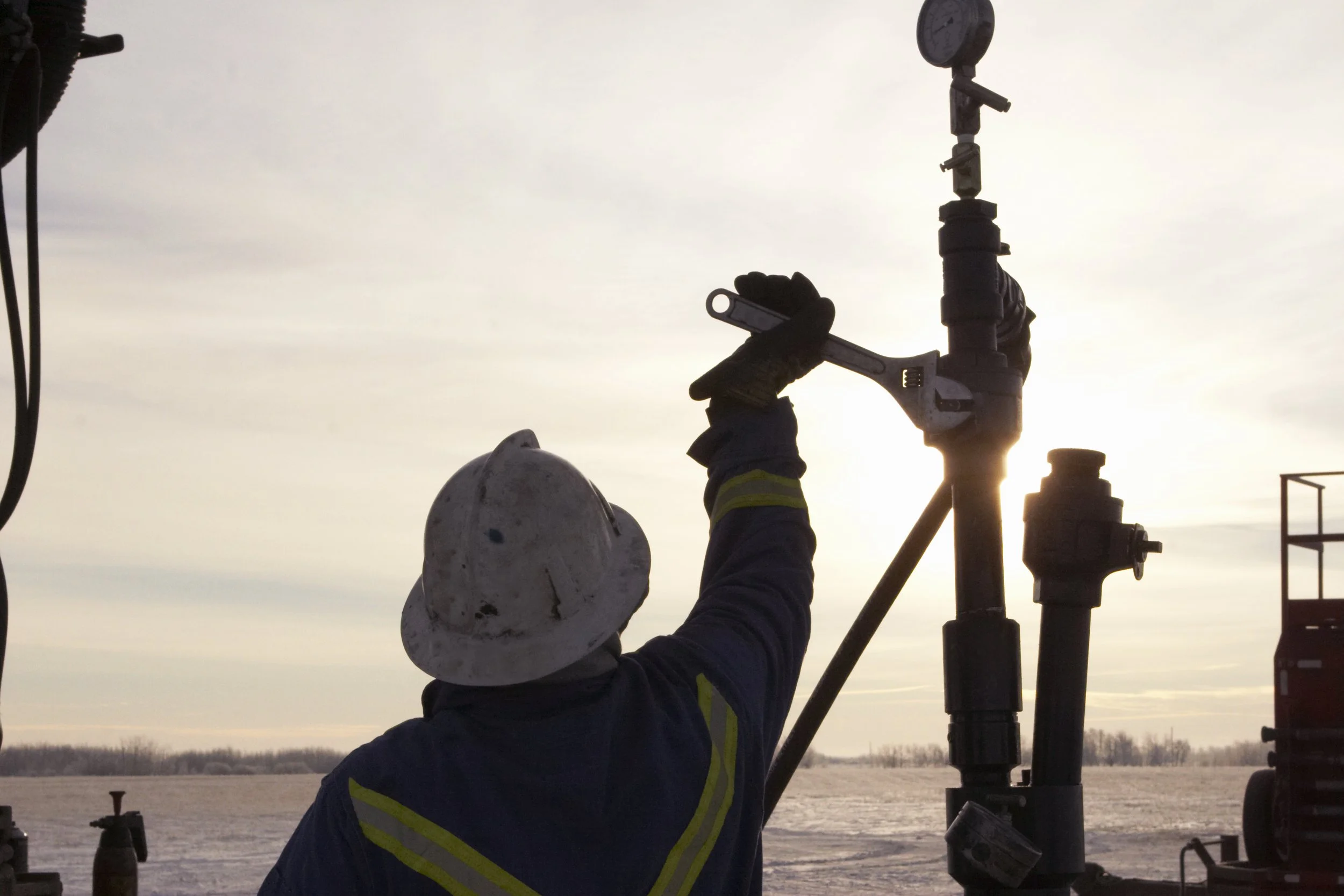 A worker outdoors at sunrise or sunset, wearing a white safety helmet and gloves, is holding a piece of equipment connected to a pipe or valve.