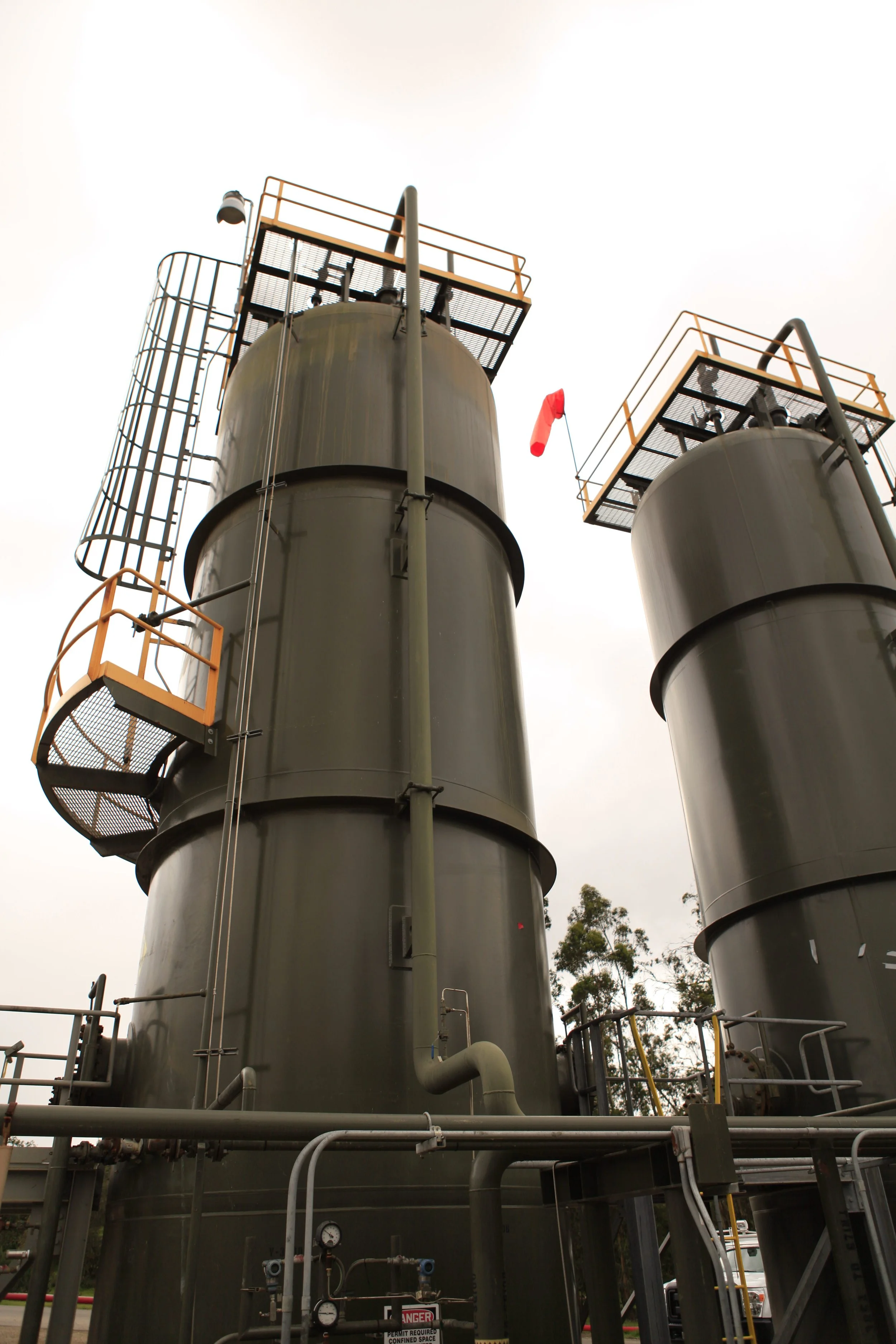Two large industrial stainless steel tanks with safety railings and ladders, a red windsock, and pipes at an industrial facility under an overcast sky, with trees in the background.