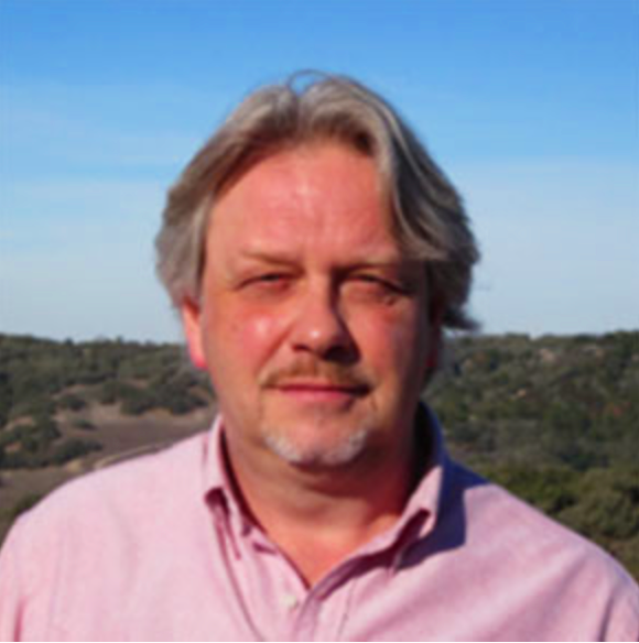 A middle-aged man with gray hair and a beard, wearing a pink collared shirt, standing outdoors with a landscape background under a blue sky.