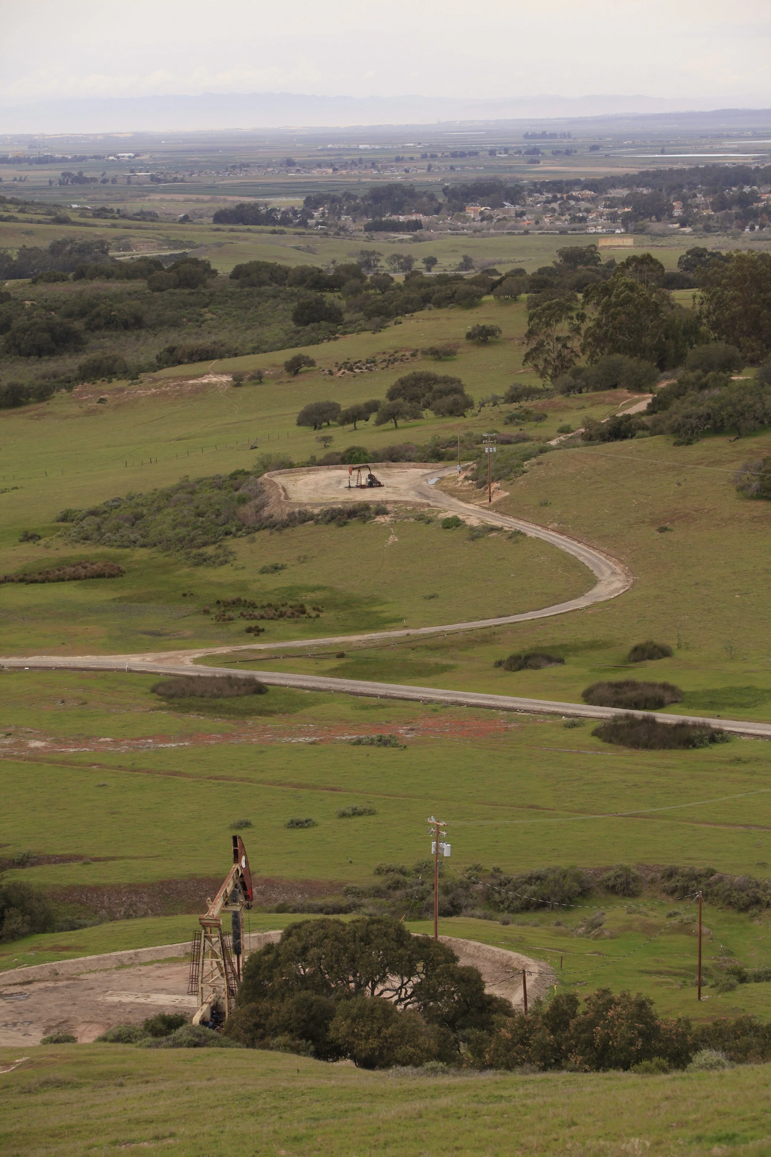 Hilly landscape with green grass, scattered trees, dirt roads, and oil pumpjacks in the foreground. Distant towns and mountains are visible under a cloudy sky.
