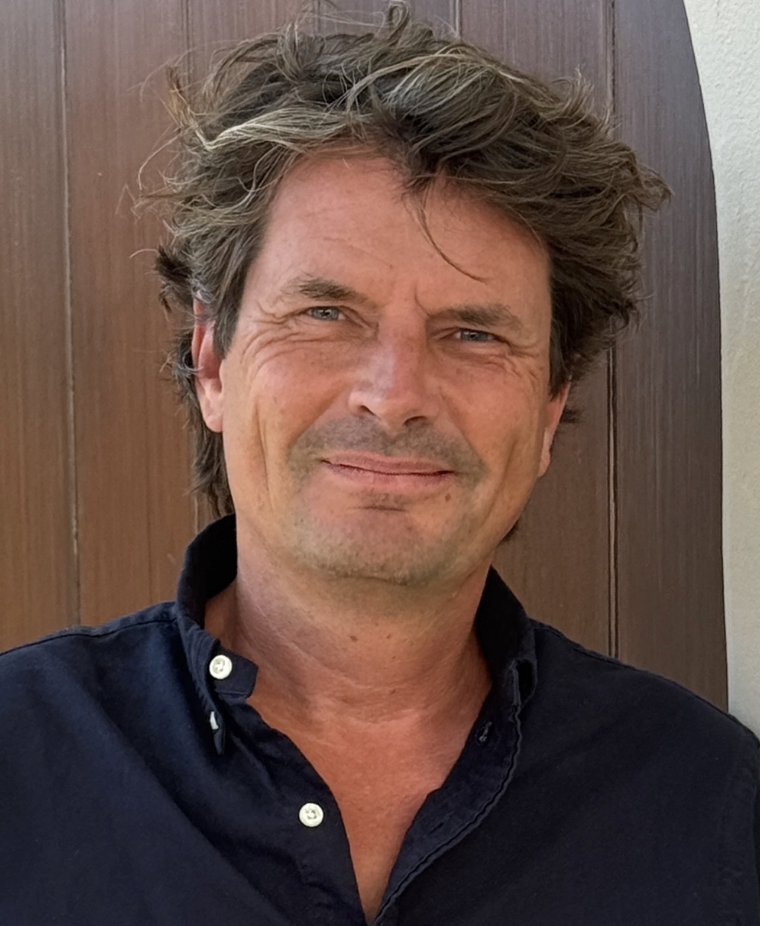 A man with wavy, tousled brown hair, wearing a dark button-up shirt, standing in front of a wooden wall.