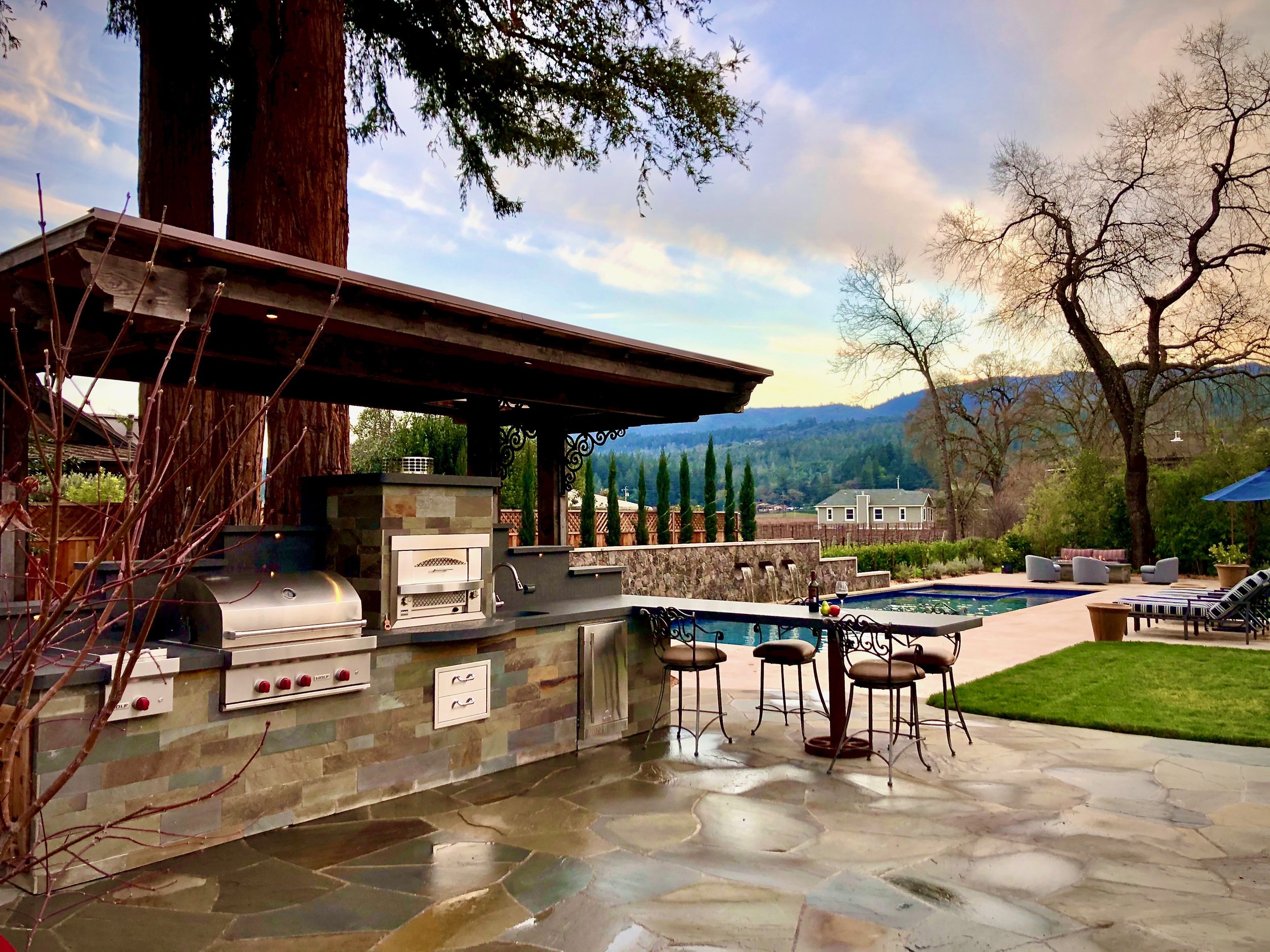 A backyard patio with a built-in grill, bar stools, and outdoor furniture, overlooking a mountain view with leafless trees and a partly cloudy sky.