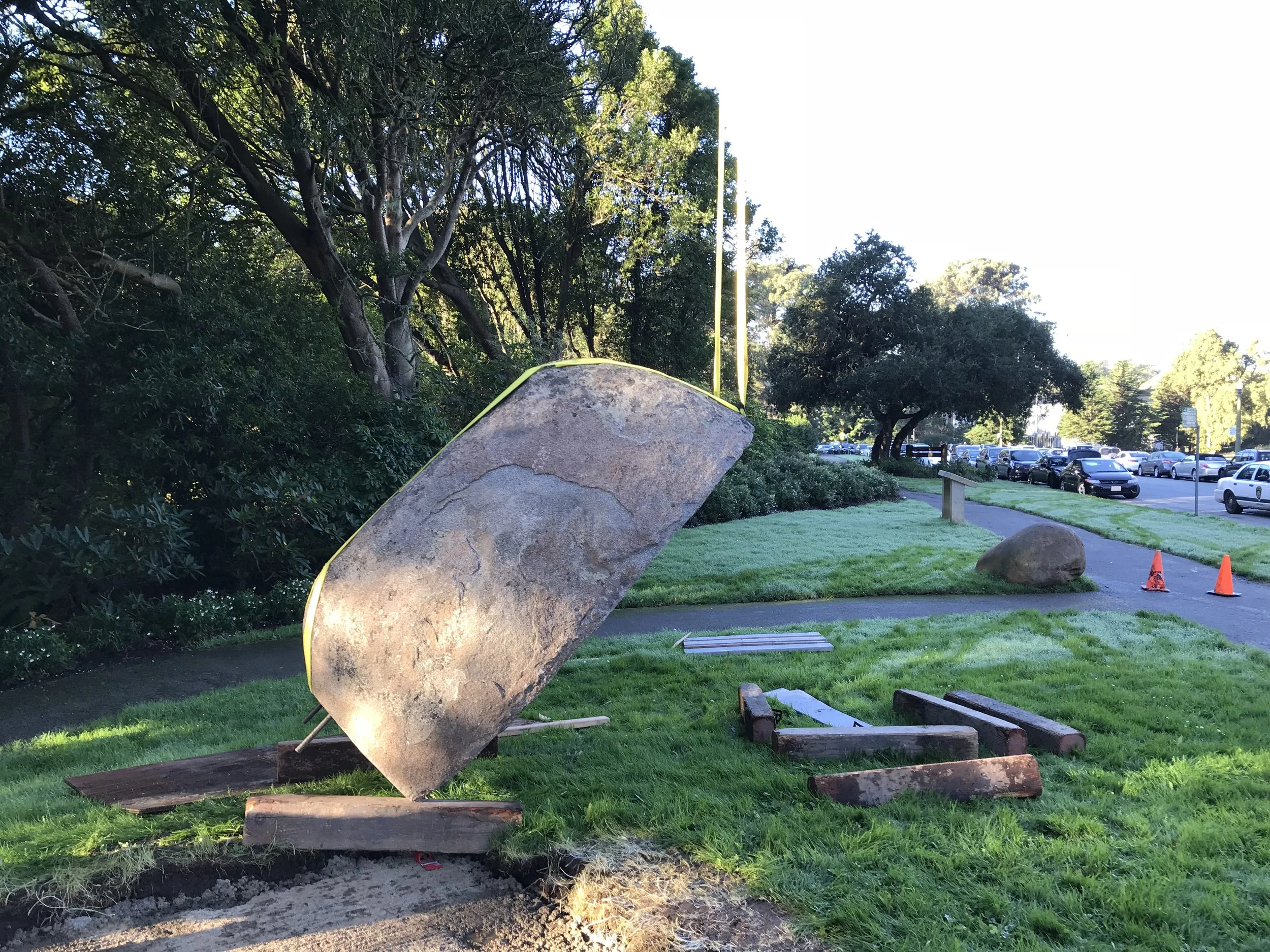 A park with large rocks and a grassy area, with trees and a parking lot in the background. Two orange traffic cones are positioned on the pavement. One prominent large rock is illuminated by sunlight.