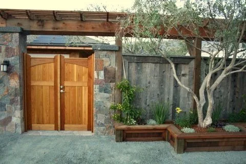 Wooden gate in a stone wall with a garden bed containing a tree and various plants.
