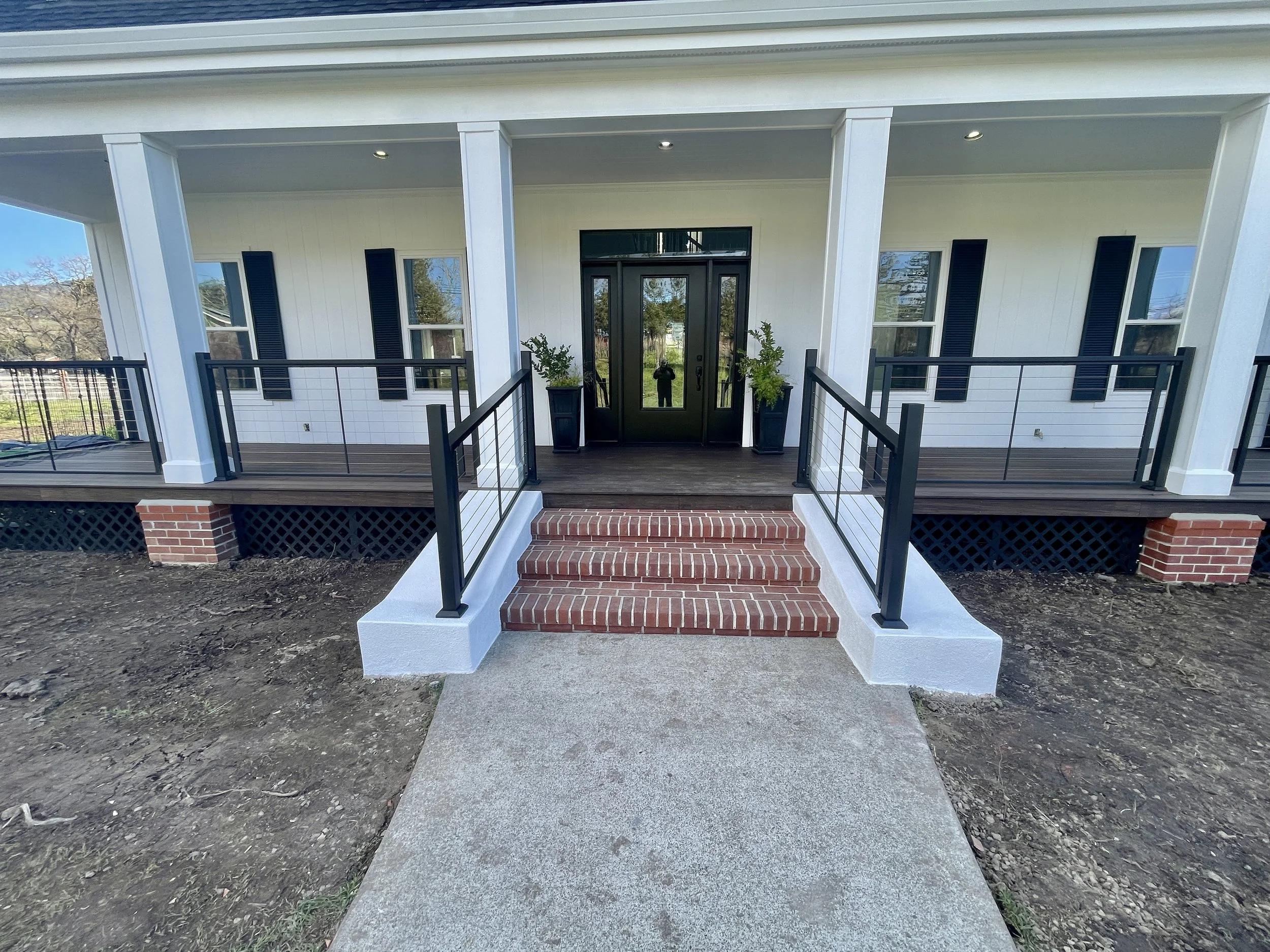 Front porch of a house with white siding, black door, black window shutters, brick steps, and potted plants on either side of the door. A person is reflected in the glass door. The porch has a black railing and a concrete walkway leading up to it.