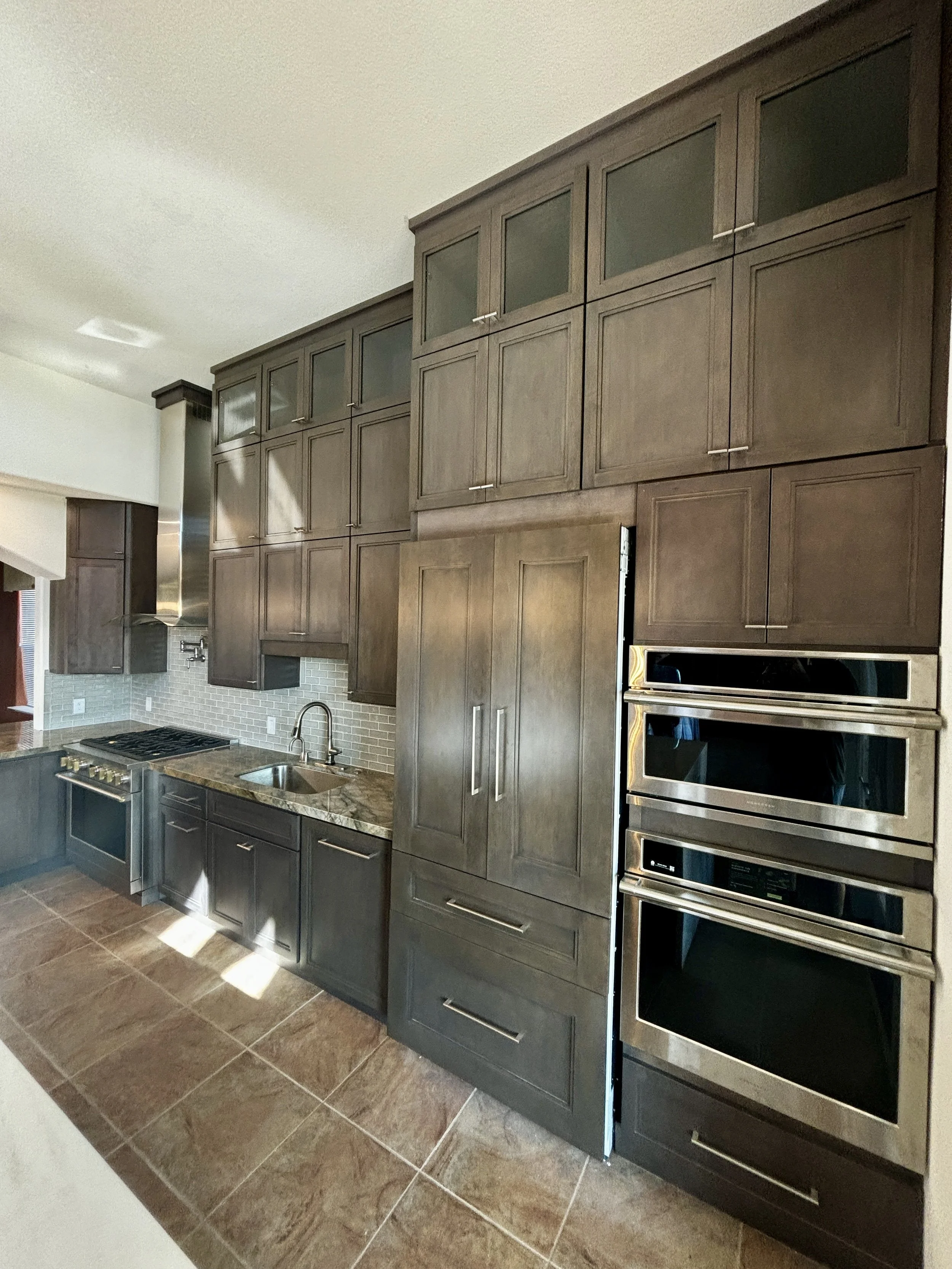 Modern remodeled kitchen with dark wood cabinets, stainless steel appliances, marble countertops, tile backsplash, and brown tiled floor.