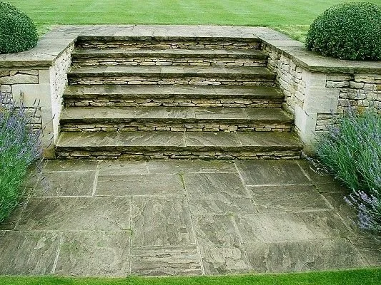 Stone steps leading uphill, flanked by shrubbery and low stone walls, in a garden setting.