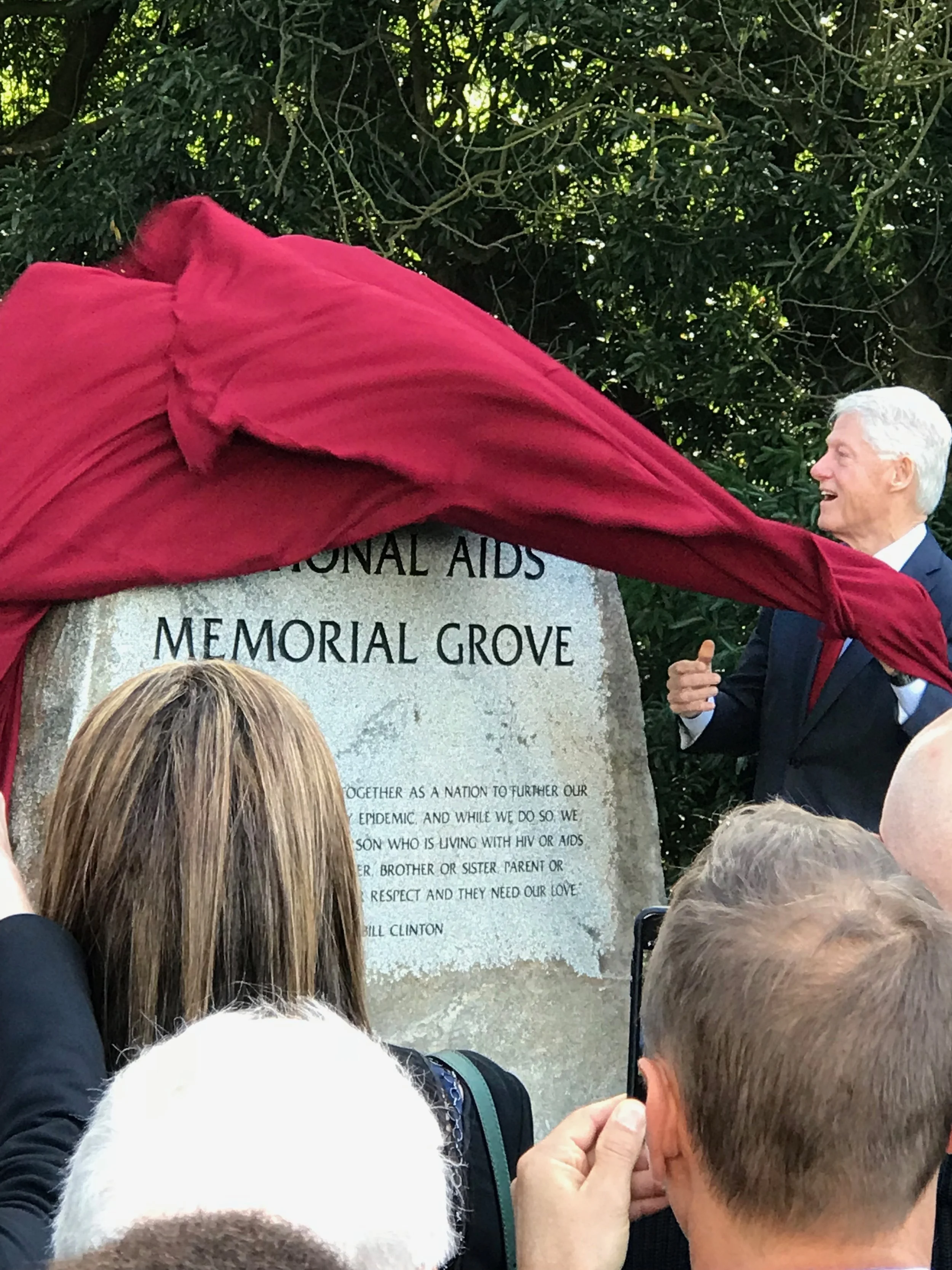 People gathered around a memorial stone with a red cloth being pulled back to reveal it, during a memorial event at Memorial Grove, with trees in the background and several individuals, mostly with gray or brown hair, observing.