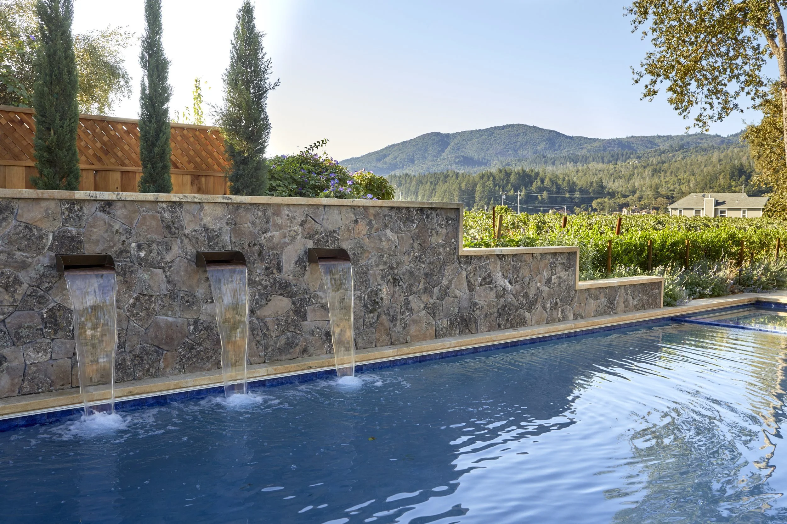 A backyard scene with a stone water feature that has three small waterfalls, a swimming pool in the foreground, a wooden privacy fence, tall cypress trees, flowering plants, green hills, and houses in the background under a clear sky.