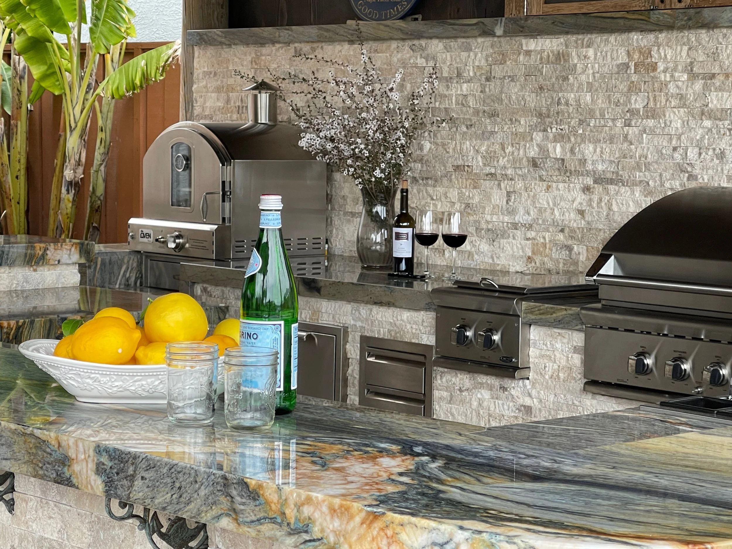 Outdoor living space with a bowl of lemons, a bottle of sparkling water, and two glasses of red wine. In the background, there is a vintage meat slicer, a large glass vase with white flowers, and a granite backsplash.